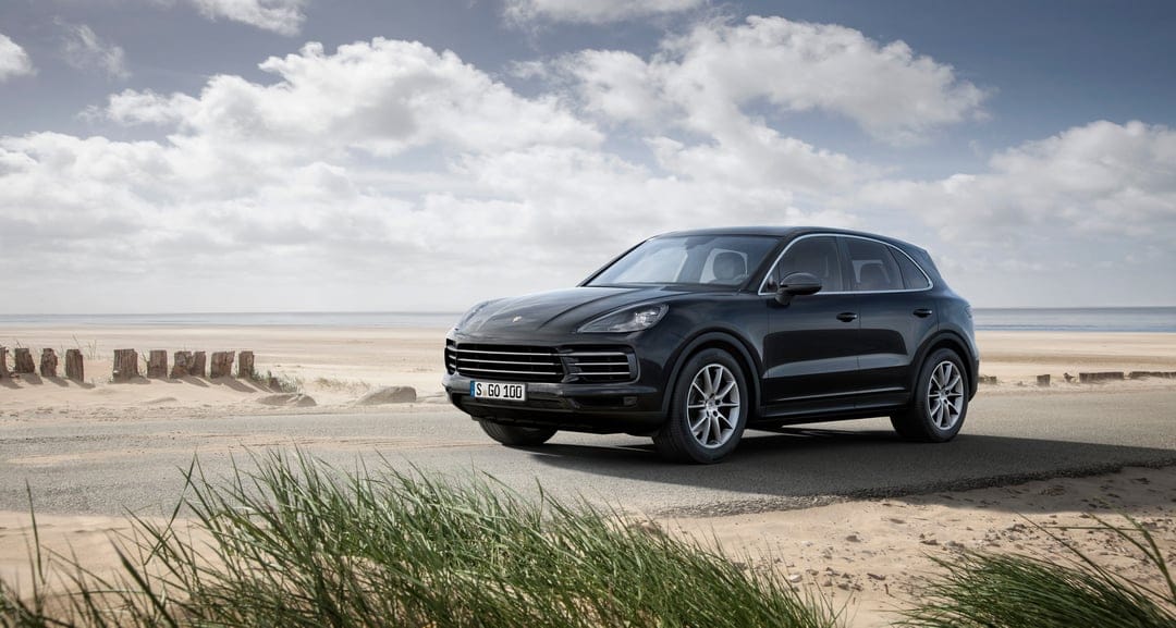 Black Porsche Cayenne SUV parked on a paved coastal road with sand dunes, beach grass, and a cloudy sky background.
