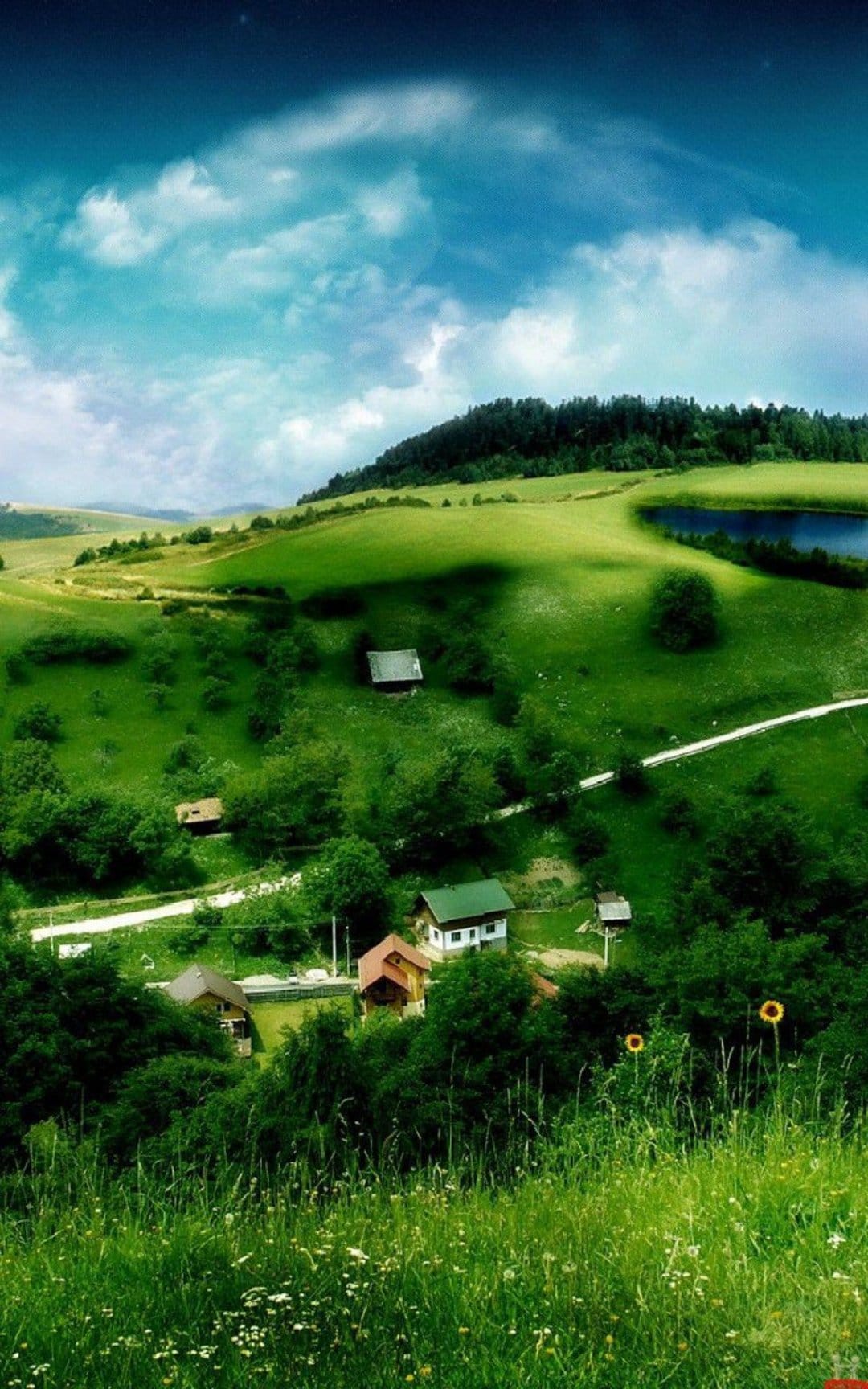 Rolling green hills with small white houses lead toward a calm lake under a bright blue sky with white clouds.