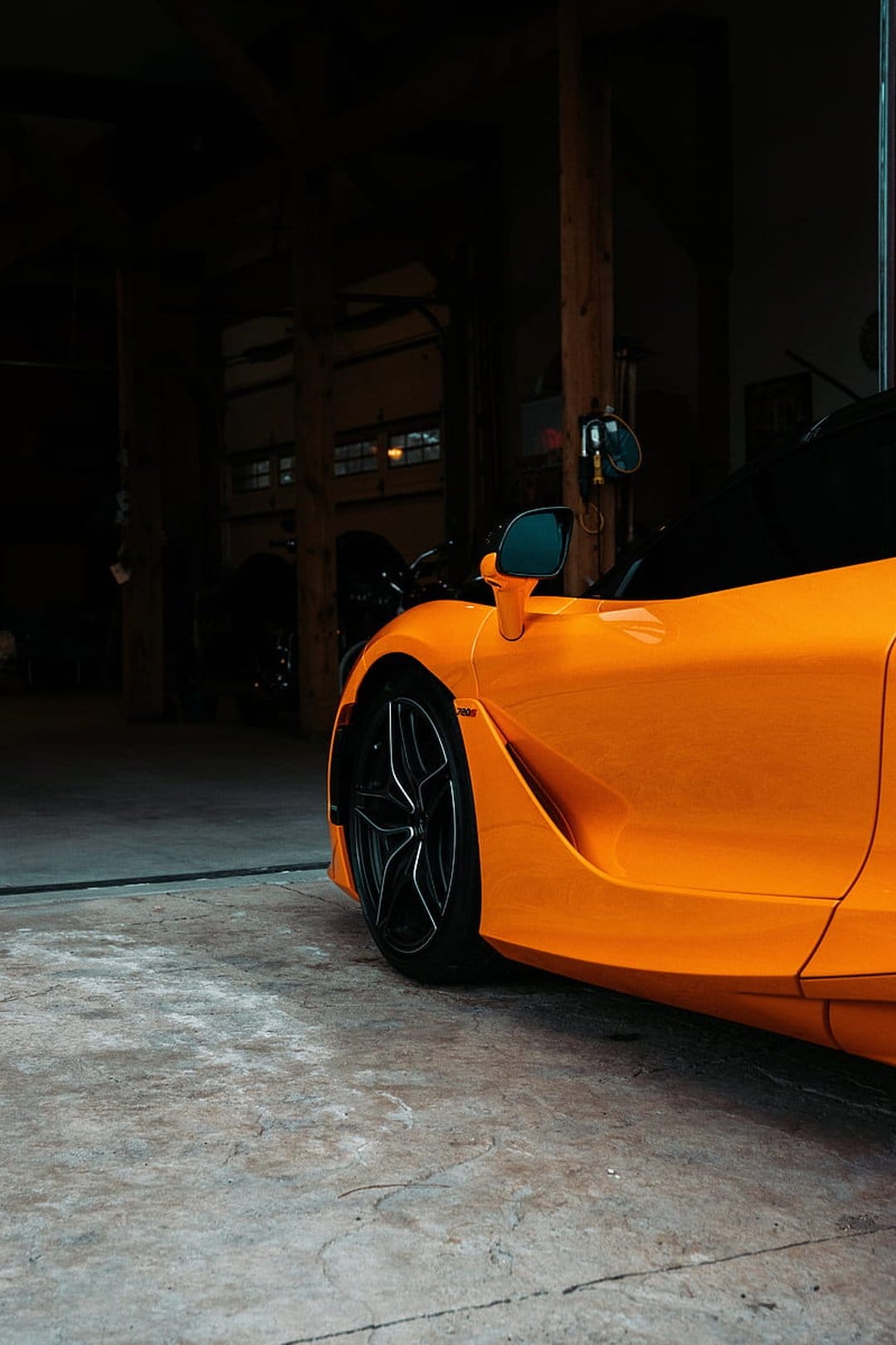 Orange McLaren sports car showing a side profile and alloy wheels in a dimly lit rustic garage setting.