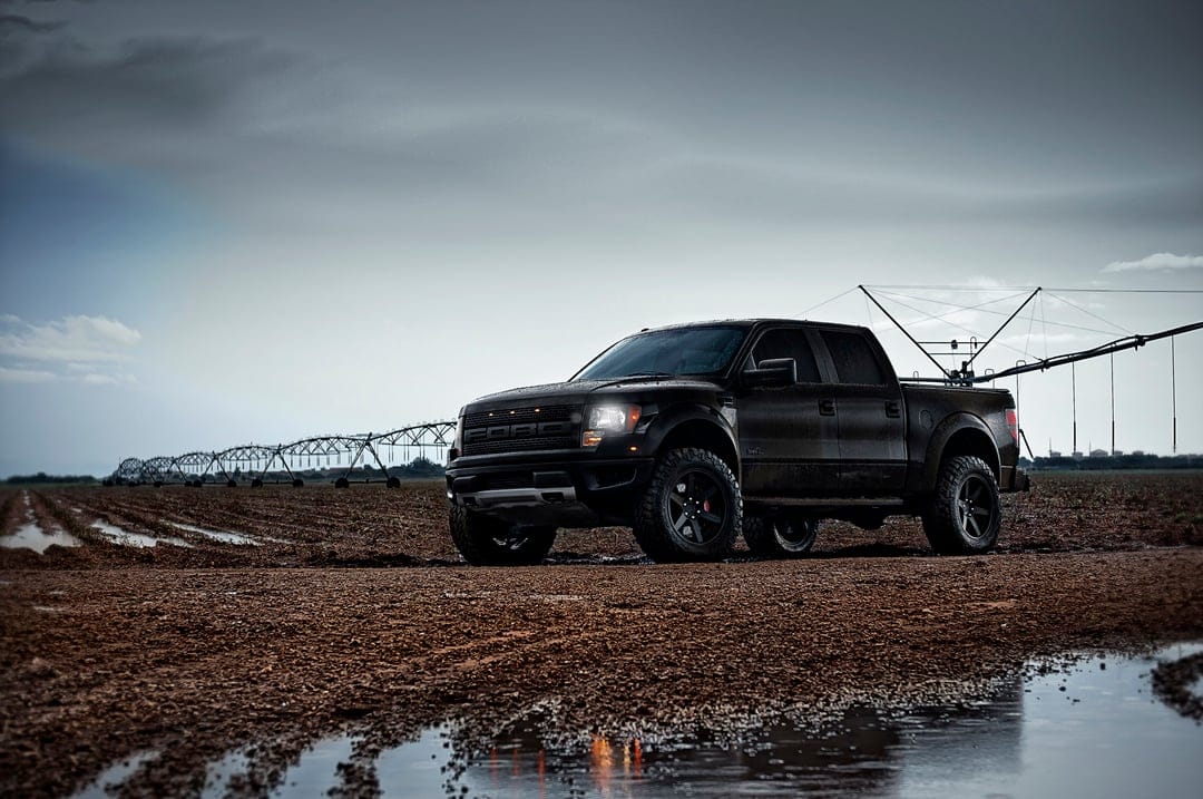 Black Ford Raptor pickup truck parked on a muddy road in a field under a dark, dramatic overcast sky.