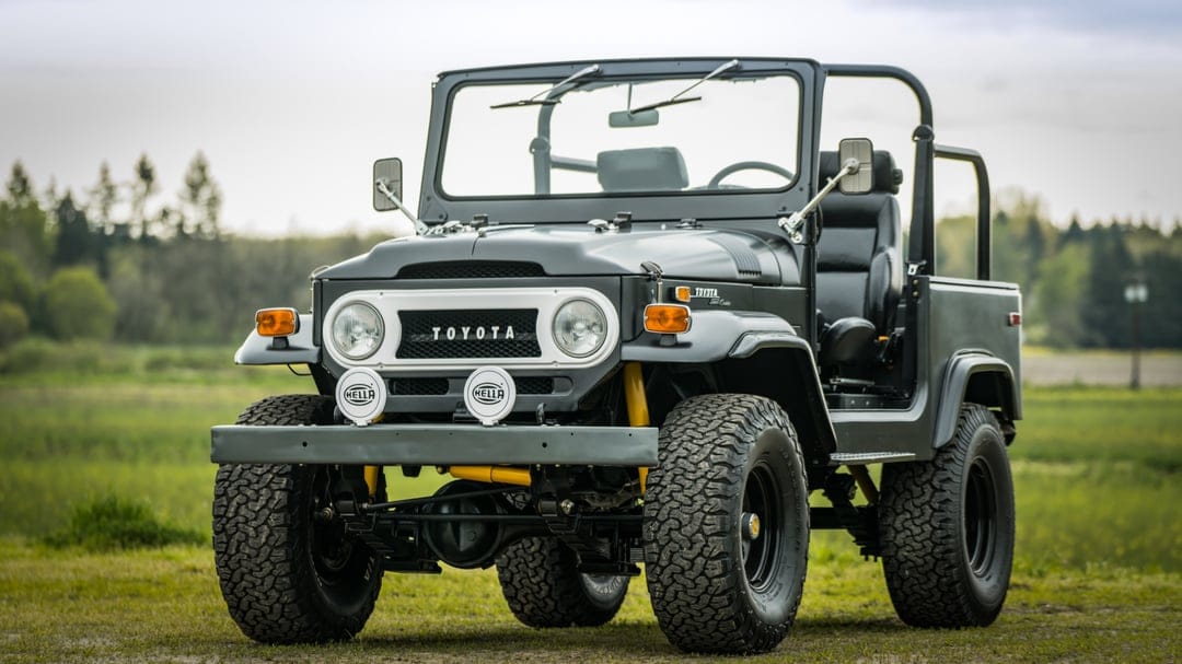 Charcoal grey Toyota Land Cruiser with off-road tires and Hella lights parked in a green field under a soft sky.