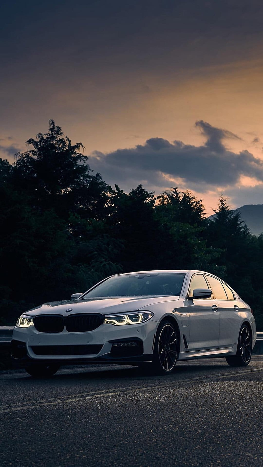 White BMW sedan with glowing headlights parked on a mountain road during a cloudy golden hour sunset.