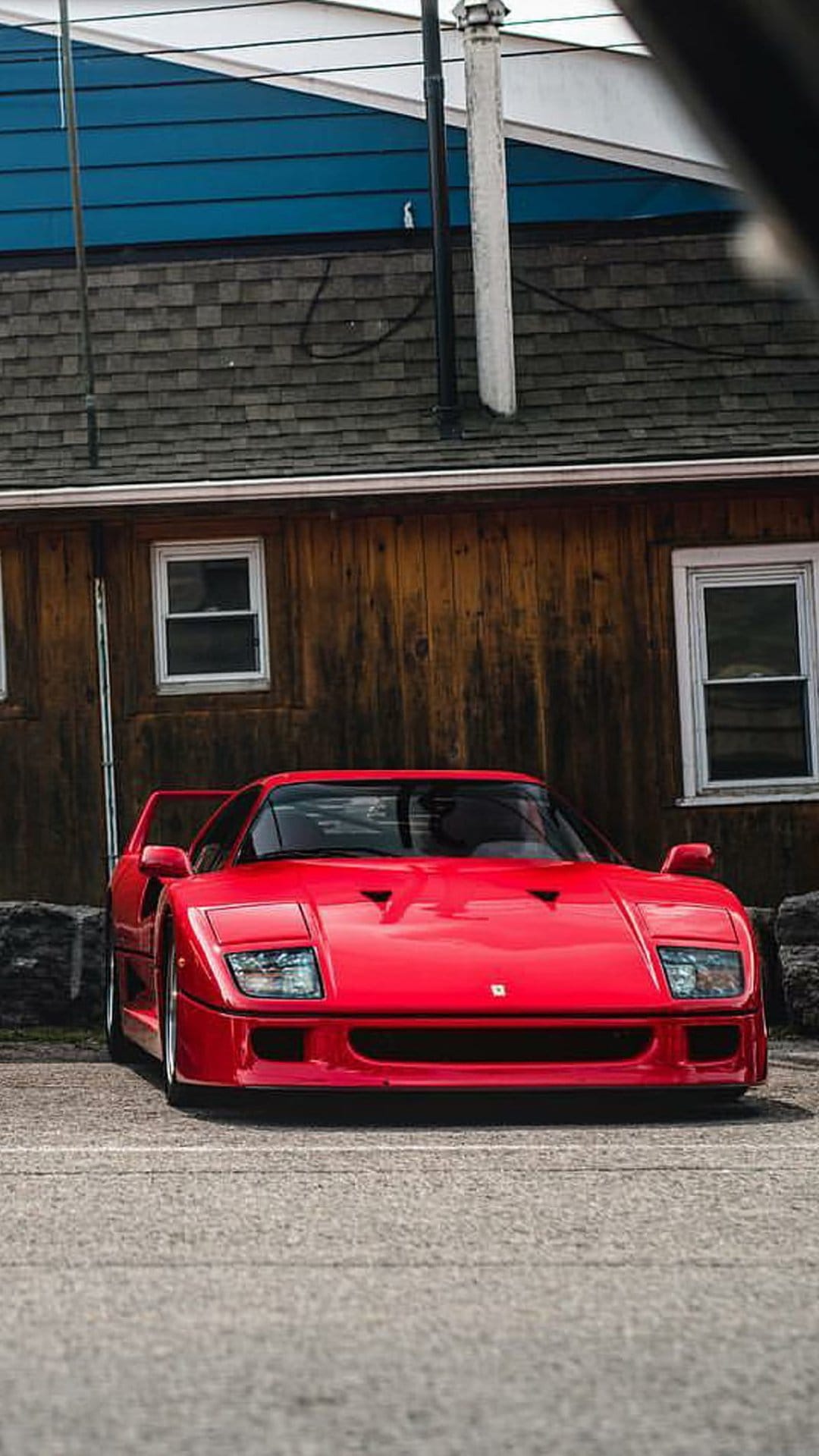 Red Ferrari F40 sports car parked outside a weathered wooden building with blue panels and rustic textures.