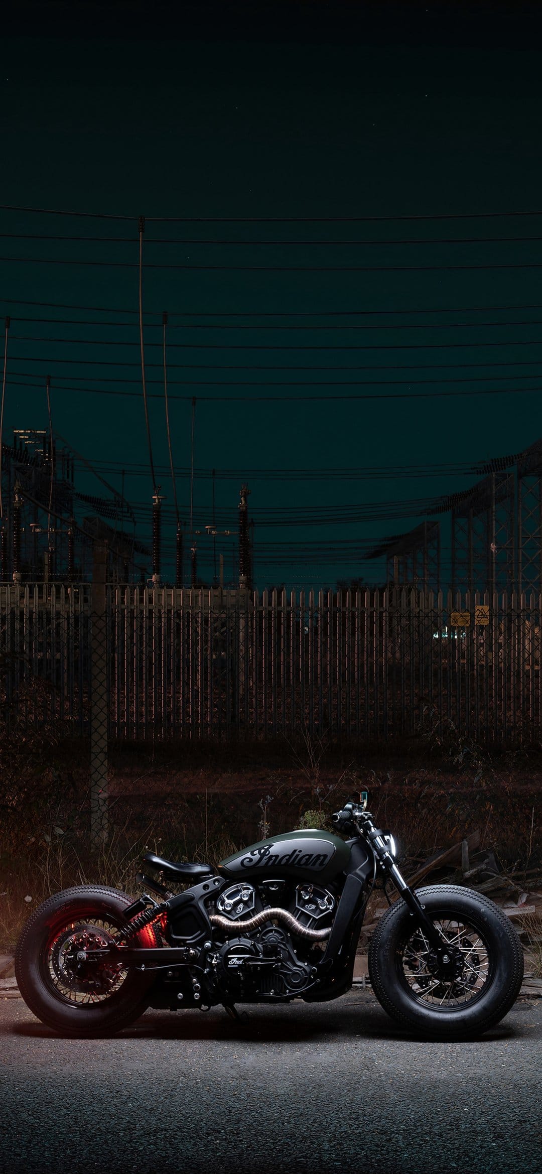 Black Indian motorcycle parked in a dark industrial zone at night with a glowing red taillight and power lines.