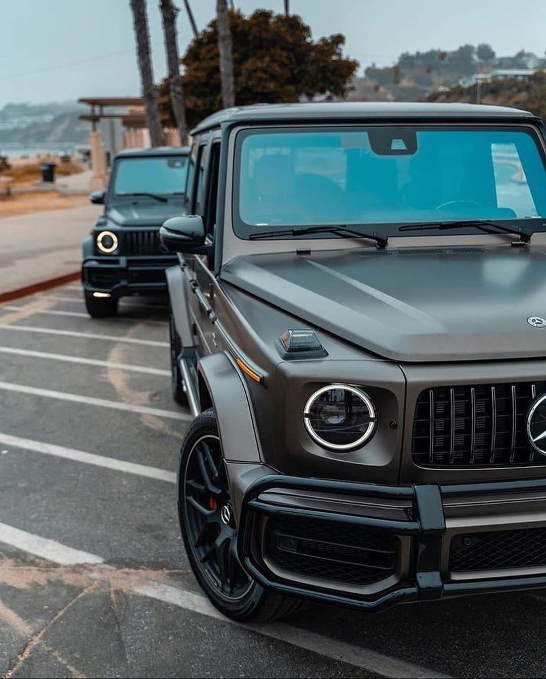 Two dark grey Mercedes G-Wagons parked on a coastal lot near palm trees and the ocean under a grey sky.