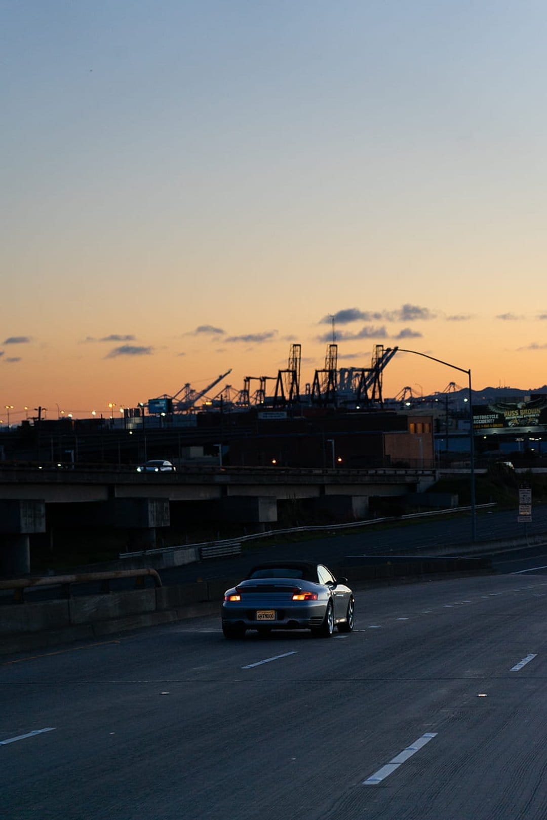 Convertible car driving on a highway at sunset with industrial port cranes silhouetted against a pink sky.