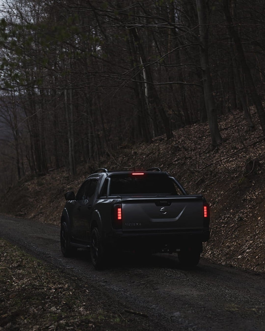 Dark pickup truck with glowing red taillights driving on a gravel road through a leafless forest at twilight.