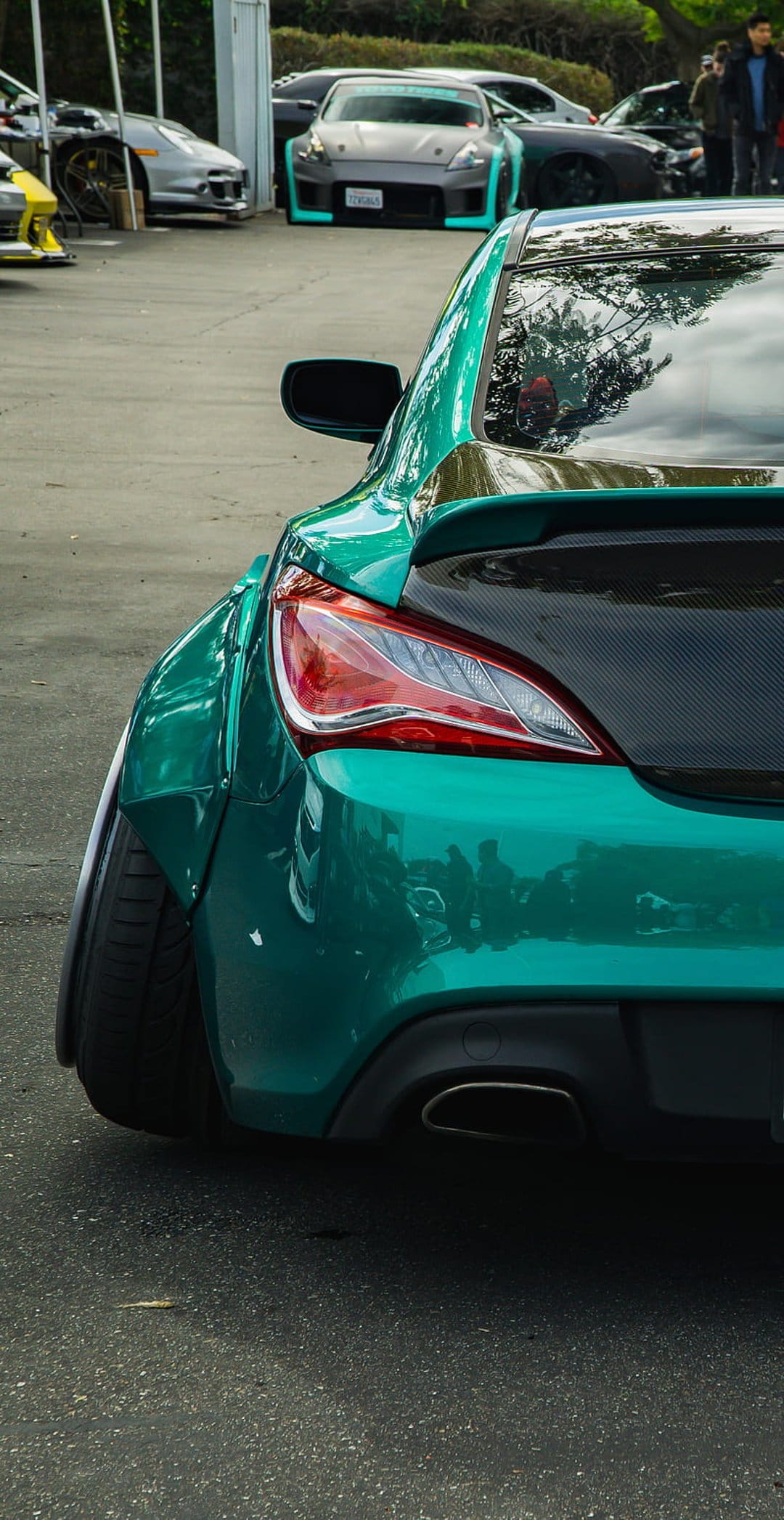 Teal sports car rear view showing widebody fenders and a carbon fiber wing at an outdoor car exhibition.
