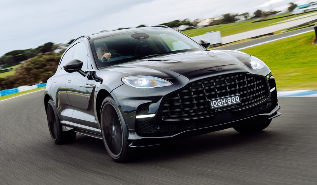 Black Aston Martin SUV driving fast on a race track with motion-blurred wheels and green grassy background.