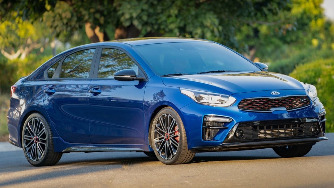 Blue Kia Forte GT sedan on an asphalt road with sunlit green trees in the background during golden hour.