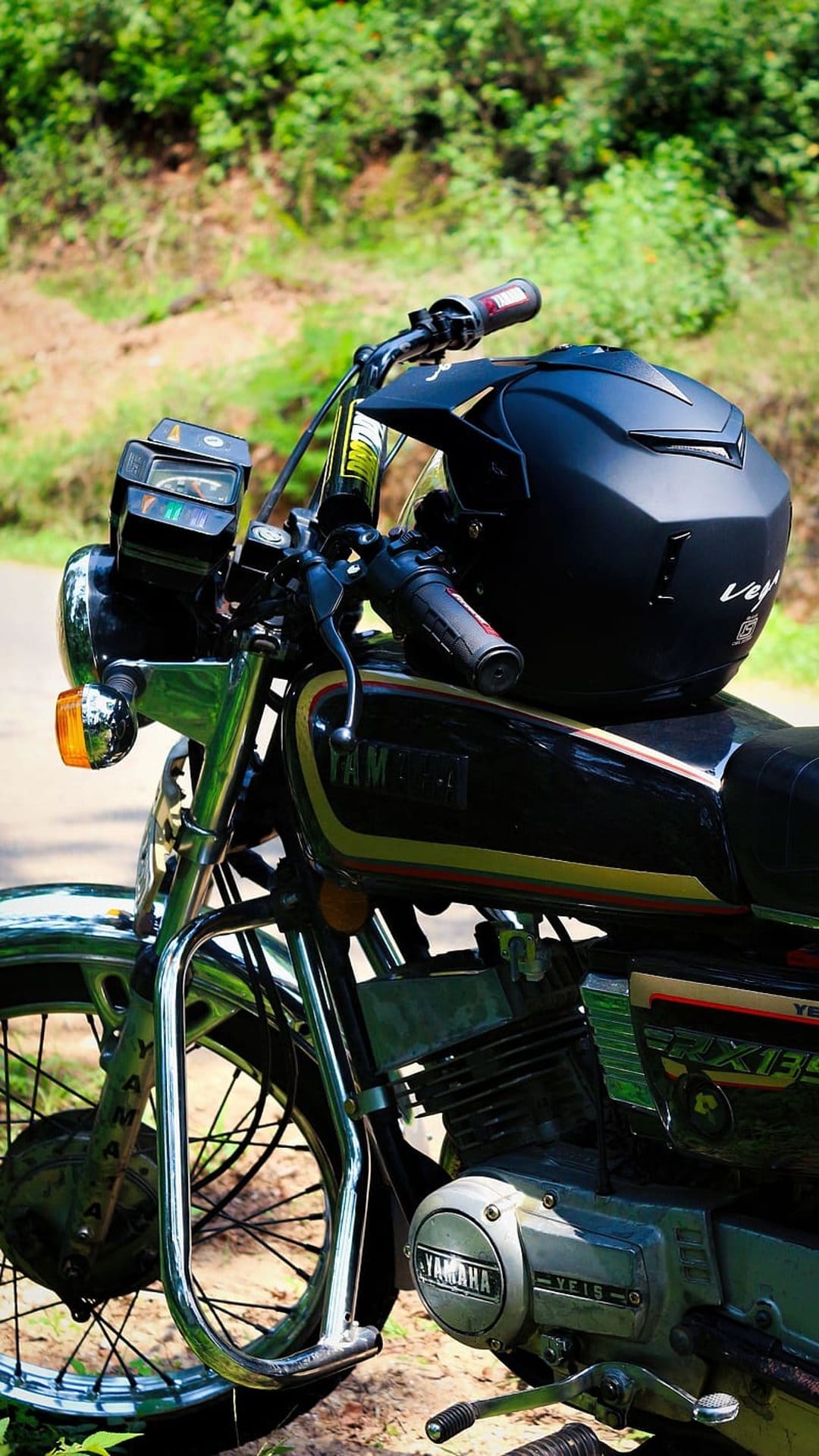 Yamaha RX 135 motorcycle with a black helmet on the seat, parked on a dirt trail surrounded by green bushes.