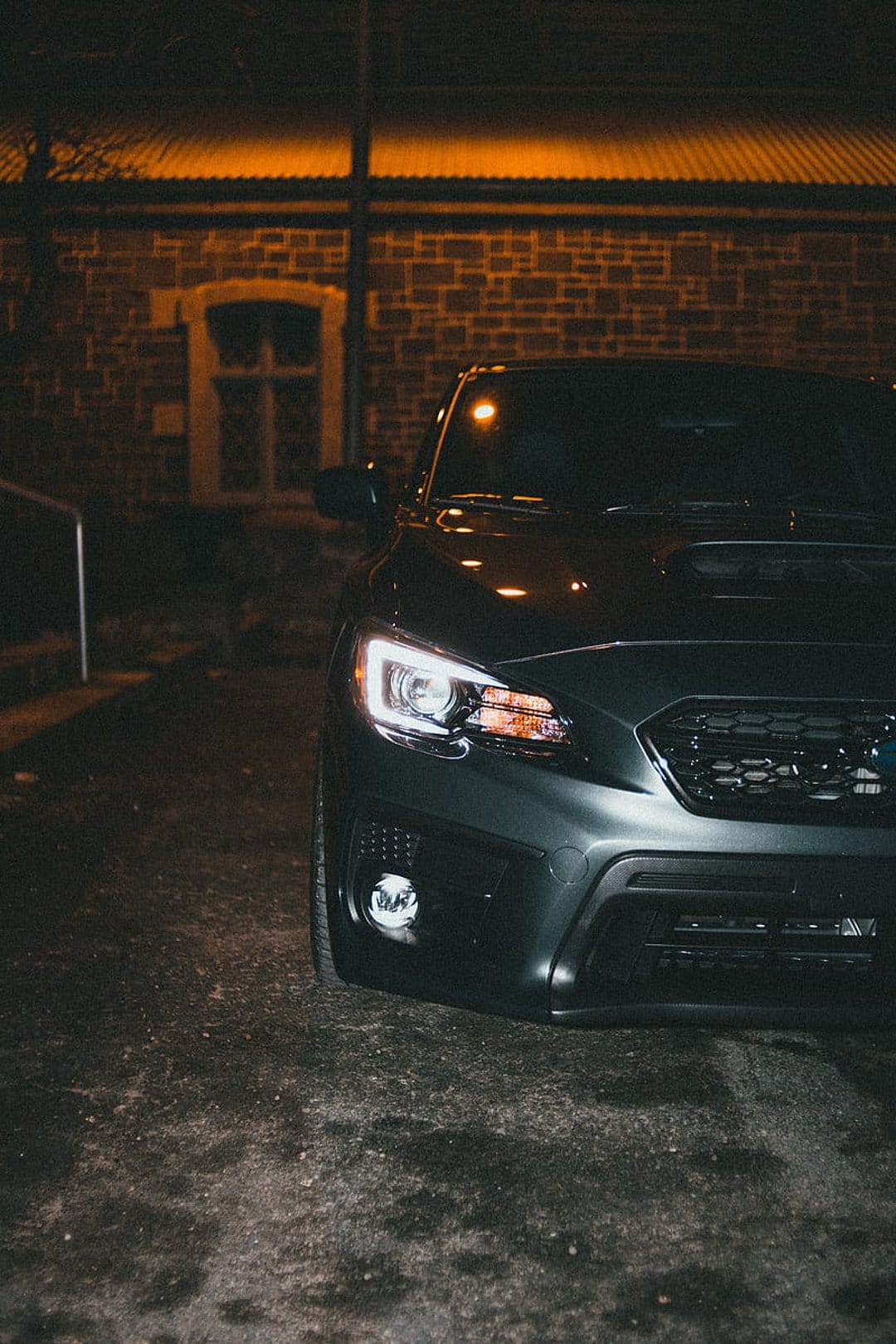 Dark Subaru WRX parked at night with headlights on against a brick building backdrop under warm orange light.