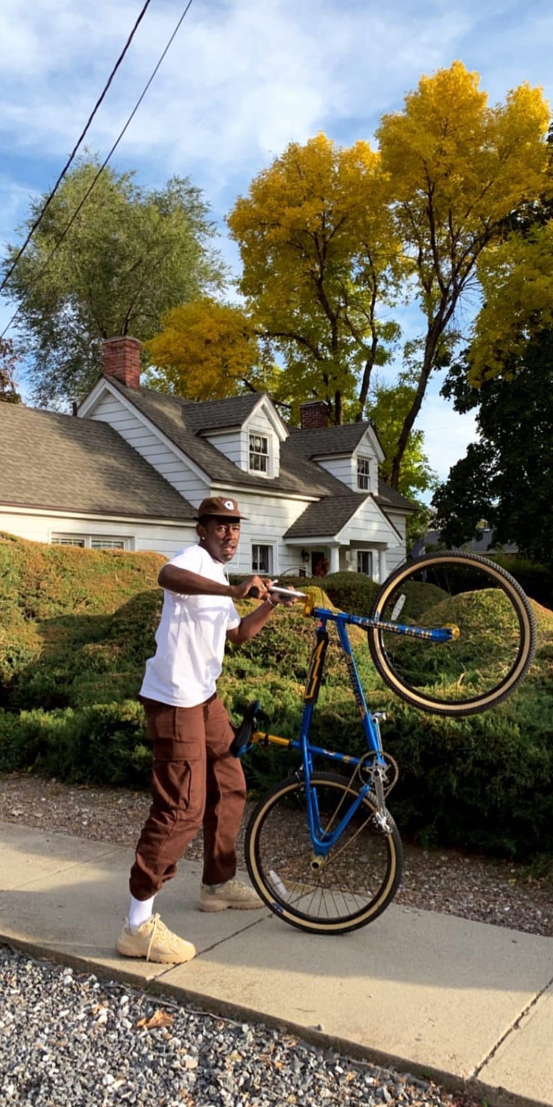Man in cargo pants lifting the front wheel of a blue bicycle on a sidewalk near yellow autumn trees and a house.