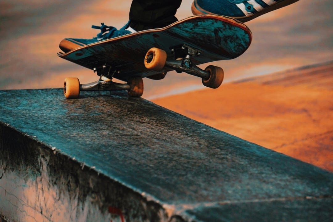 Skateboarder's feet and a teal board grind a concrete ramp edge under a blurred orange and pink sunset sky.