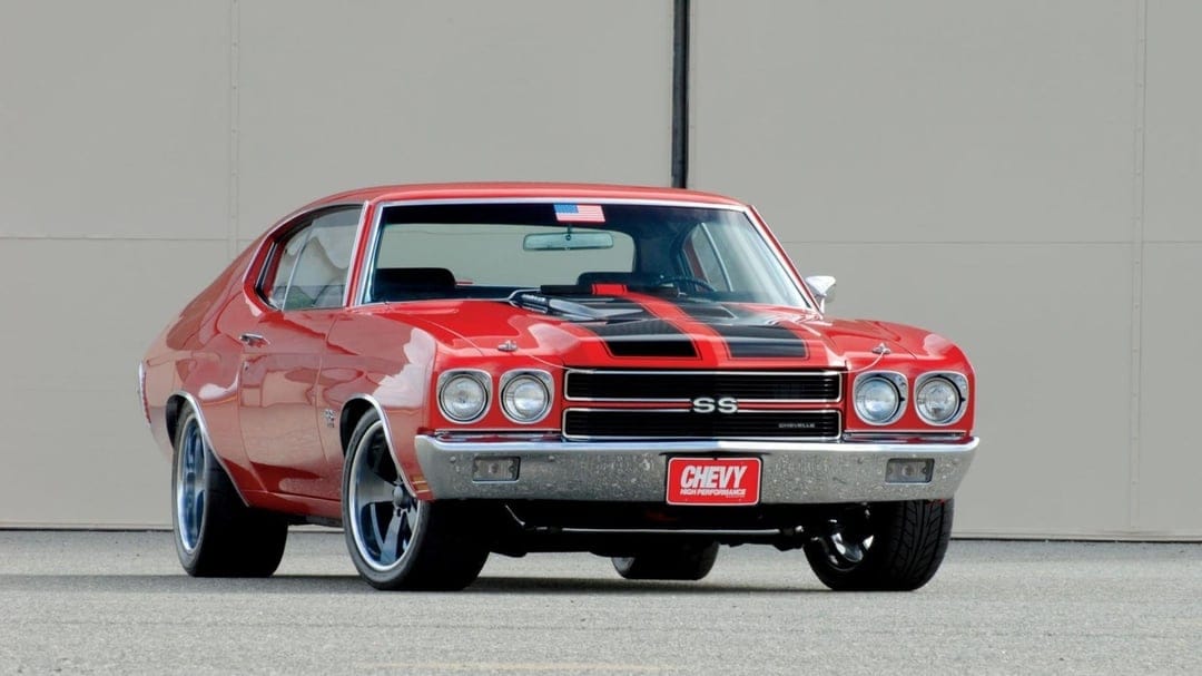 Red 1970 Chevrolet Chevelle SS with black racing stripes and chrome bumpers parked on asphalt against a tan wall.