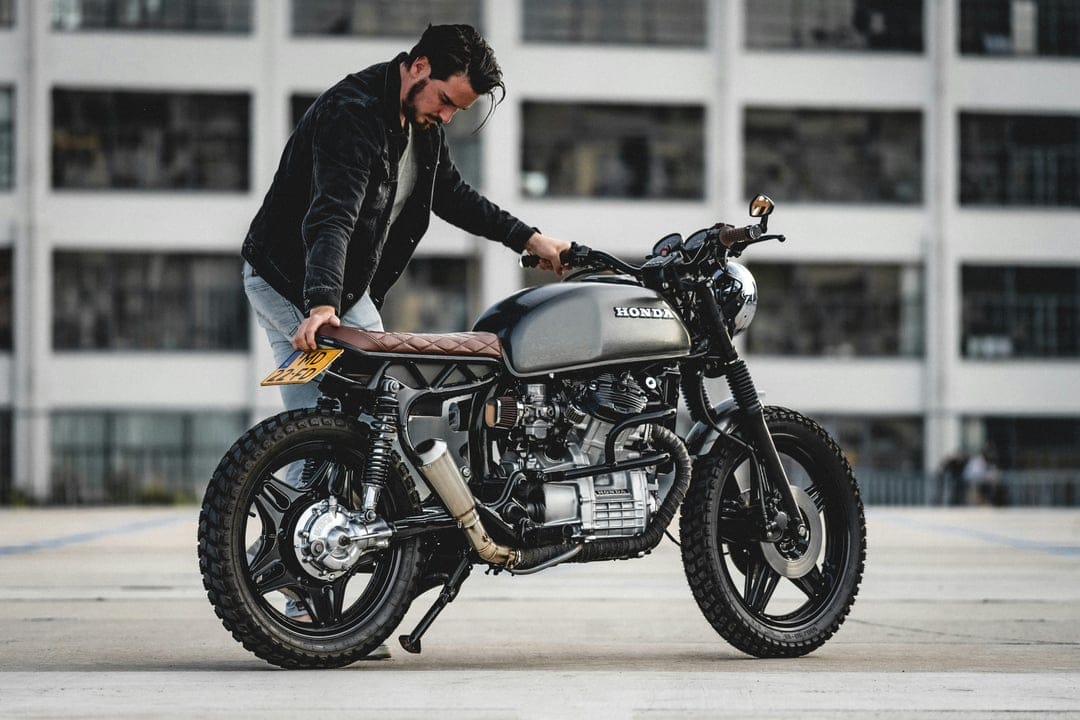 Man in a black jacket stands by a custom Honda motorcycle with a brown leather seat and grey tank in a city.