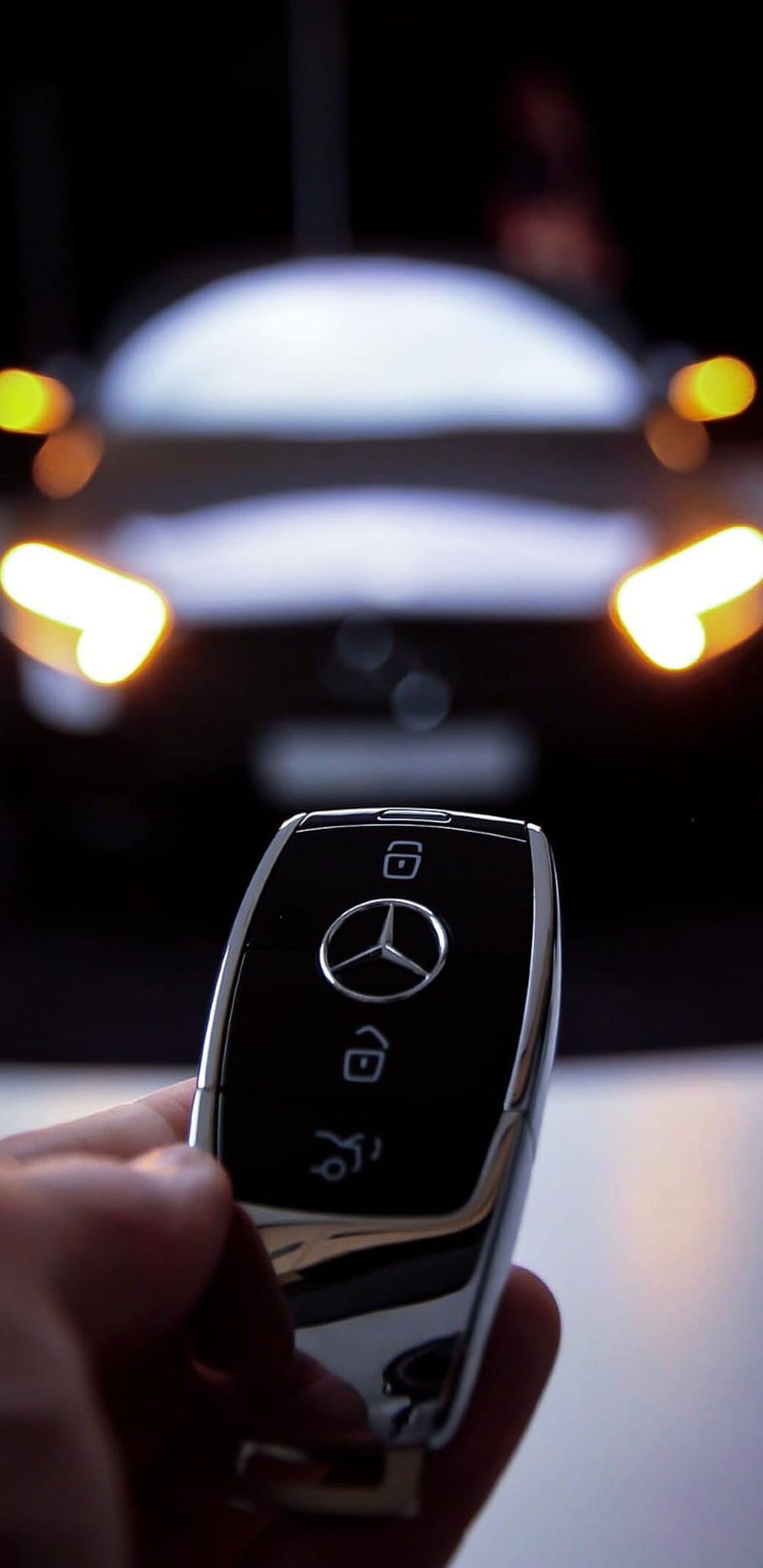 Mercedes-Benz key fob held in focus with glowing circular car headlights blurred in the dark background.