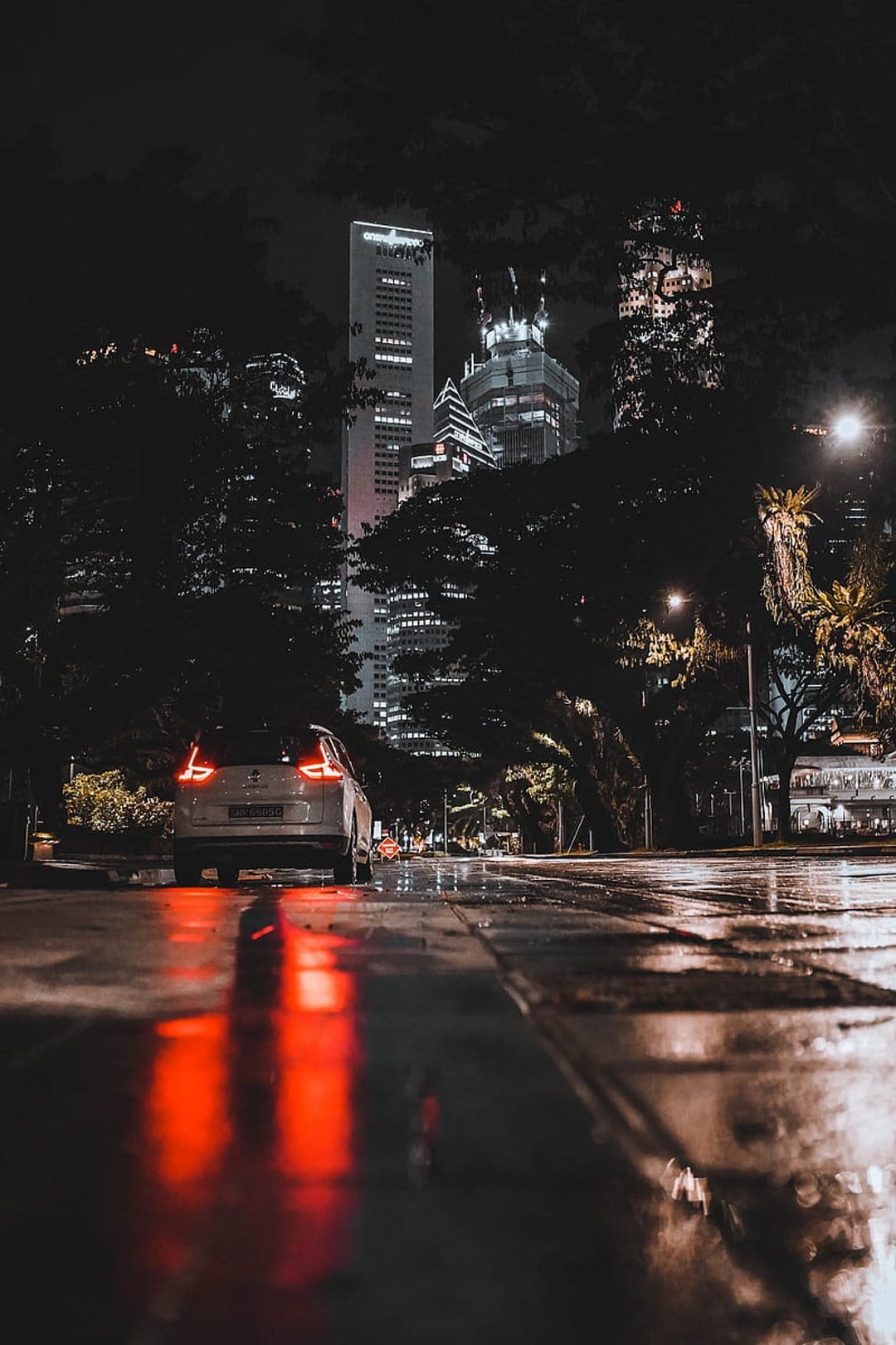 Red car taillights reflect on a wet asphalt city street lined with tall buildings under a dark rainy sky.