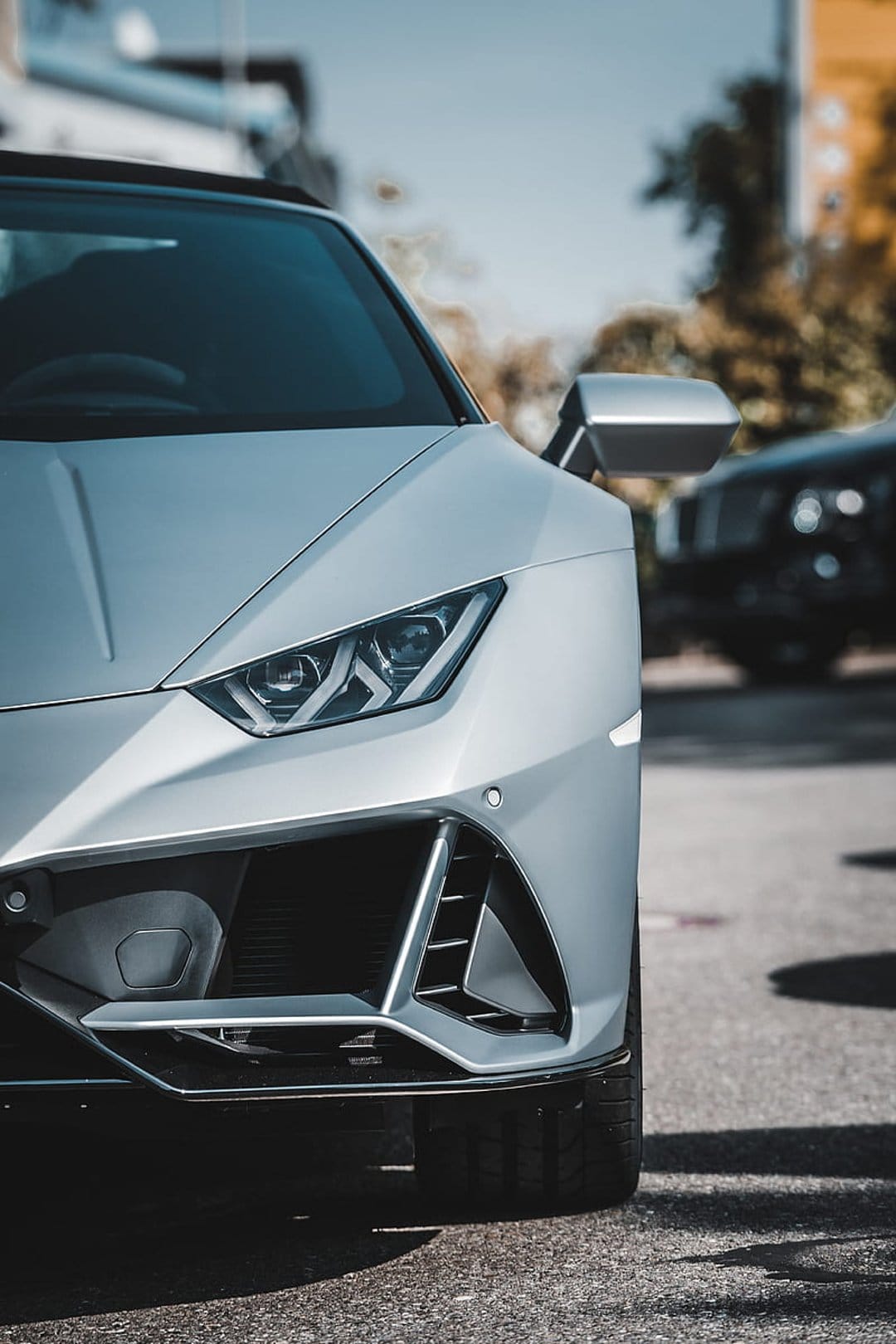 White Lamborghini Huracan front grille and LED headlights with a blurred outdoor forest background.