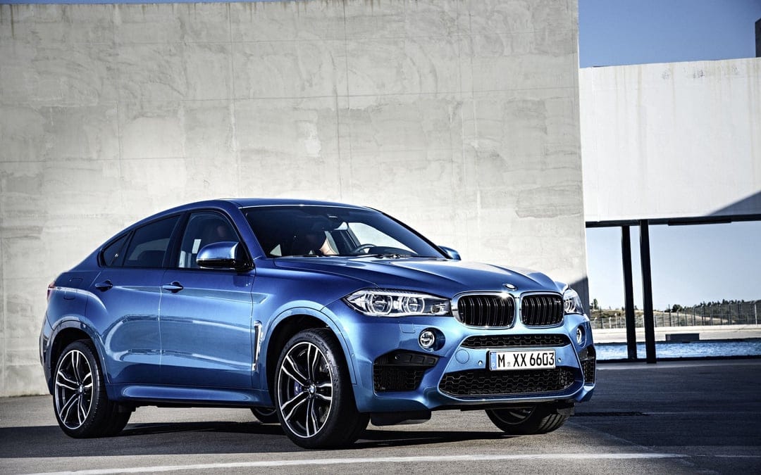 Blue BMW X6 M luxury SUV parked in front of a minimalist concrete wall with bright daylight and sharp shadows.