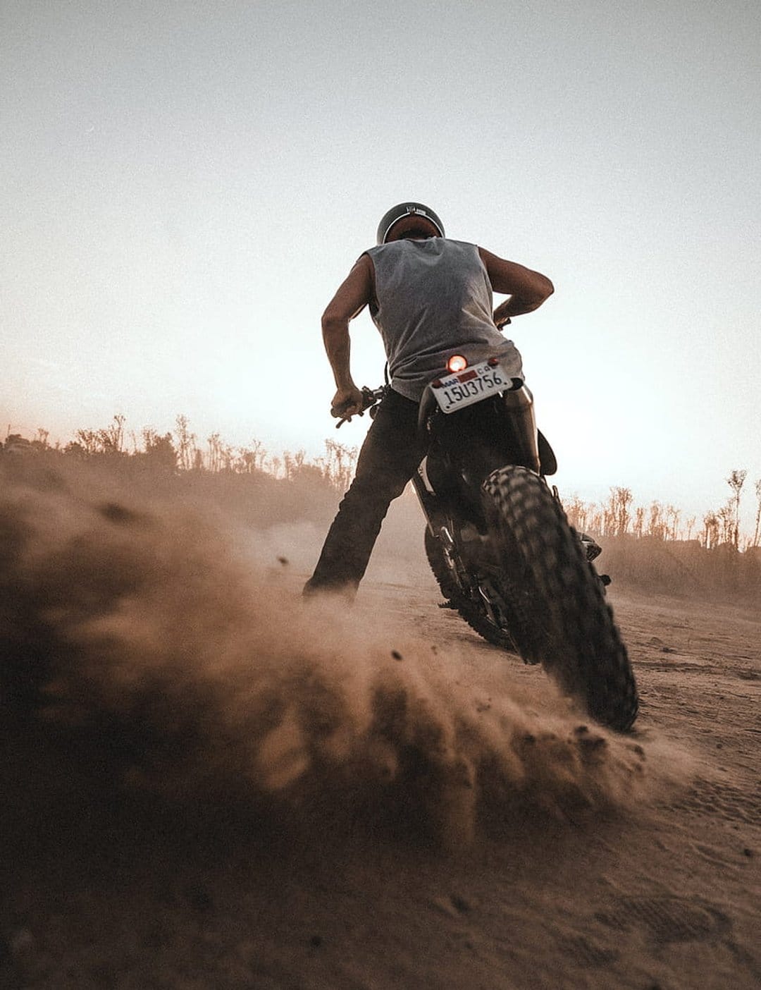 Motorcyclist in a helmet leans into a sharp turn, kicking up a massive cloud of dust on a desert track at sunset.