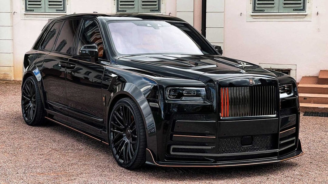 Black widebody Rolls-Royce Cullinan with orange grille accents parked on gravel against a light pink building.