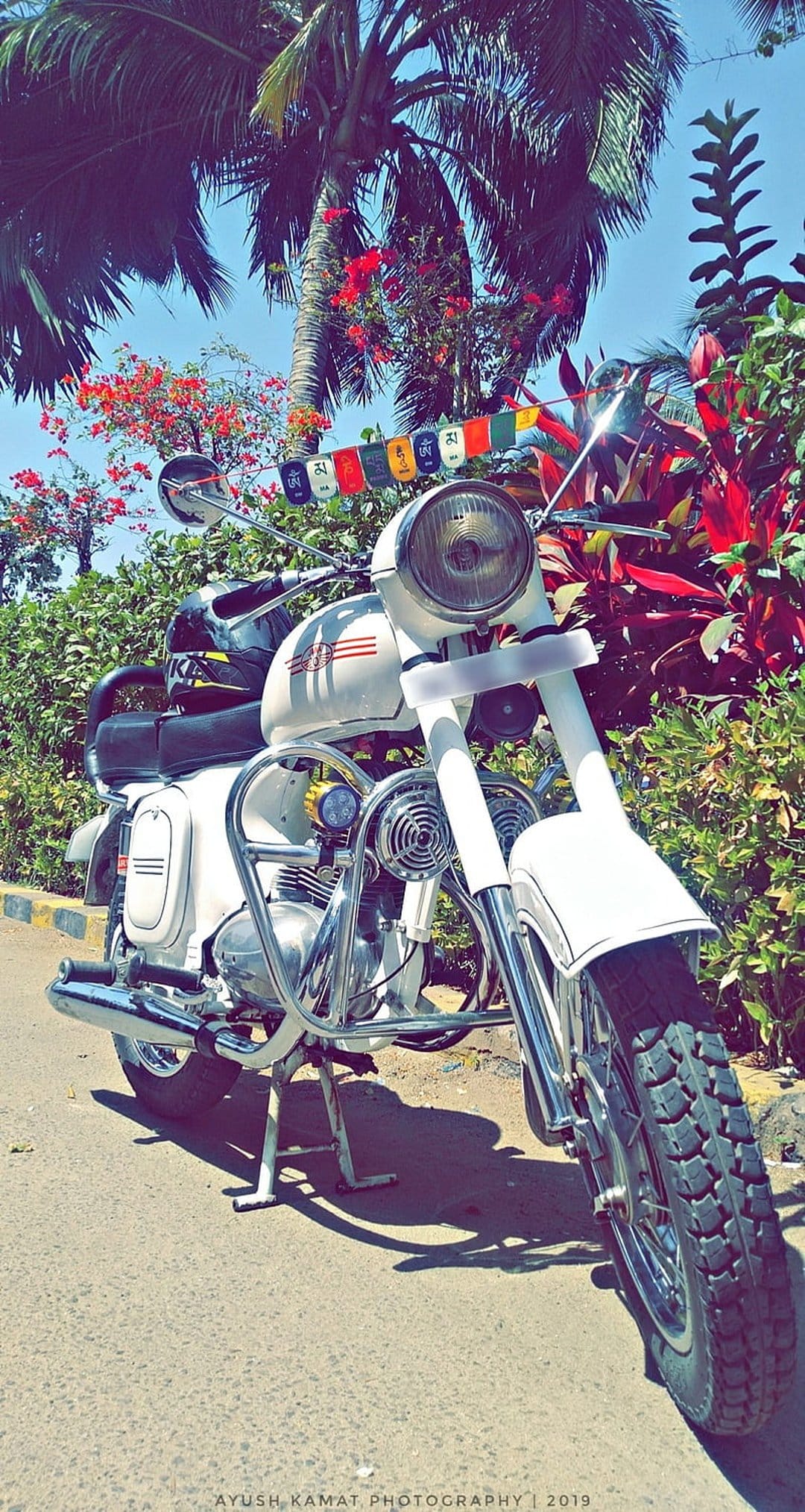 White vintage motorcycle with chrome engine parts parked among tropical palm trees and vibrant red flowers.