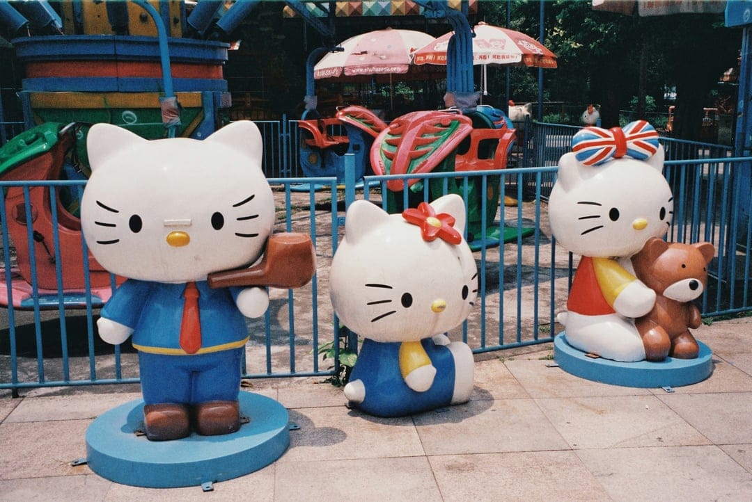 Hello Kitty statues in various outfits stand before amusement park rides and a blue fence on a sunny day.