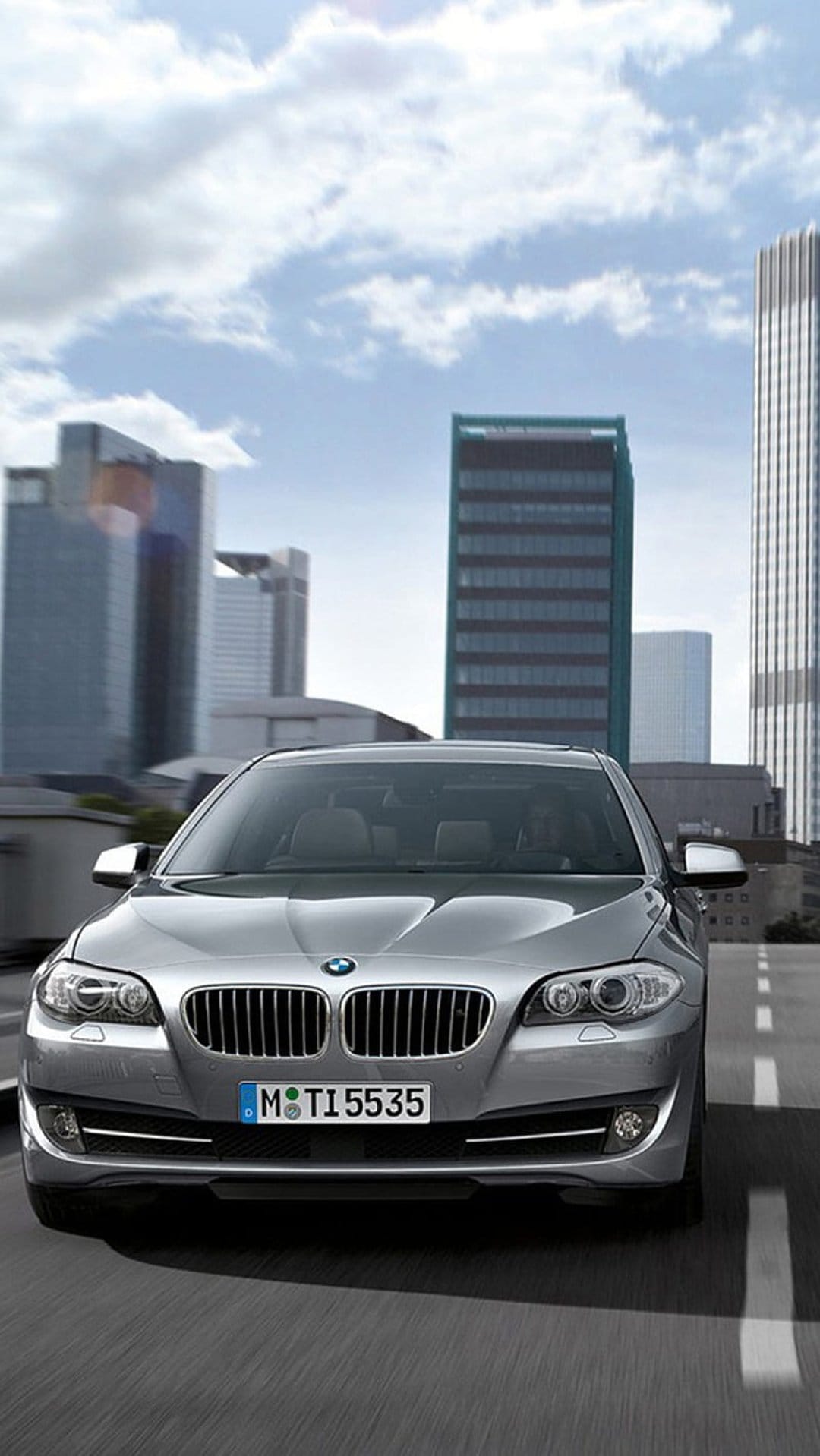 Silver BMW sedan with headlights on driving on an asphalt city road between tall glass skyscrapers and blue sky.