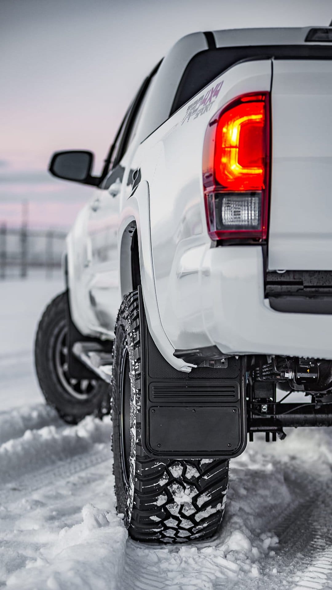 White Toyota Tacoma pickup truck with red taillights and mud flaps driving on a snowy road under an overcast sky.