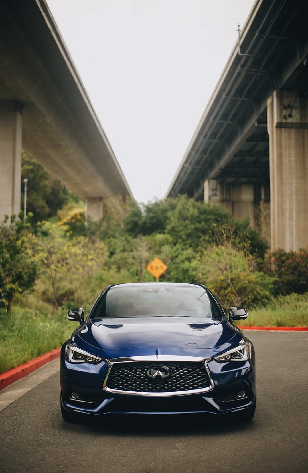 Dark blue Infiniti sedan parked on asphalt between concrete overpass supports and lush green bushes near a road sign.