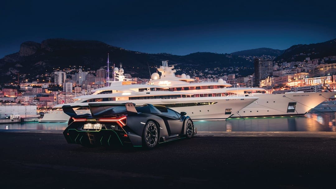 Black Lamborghini Veneno with red taillights on a pier at night by luxury yachts and glowing hillside city lights.