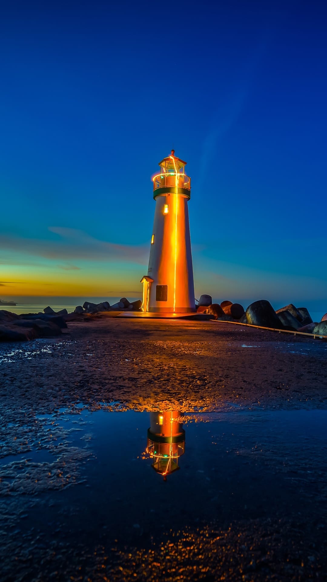 Luminous lighthouse reflecting in a ground puddle under a blue and orange twilight sky during the evening hours.