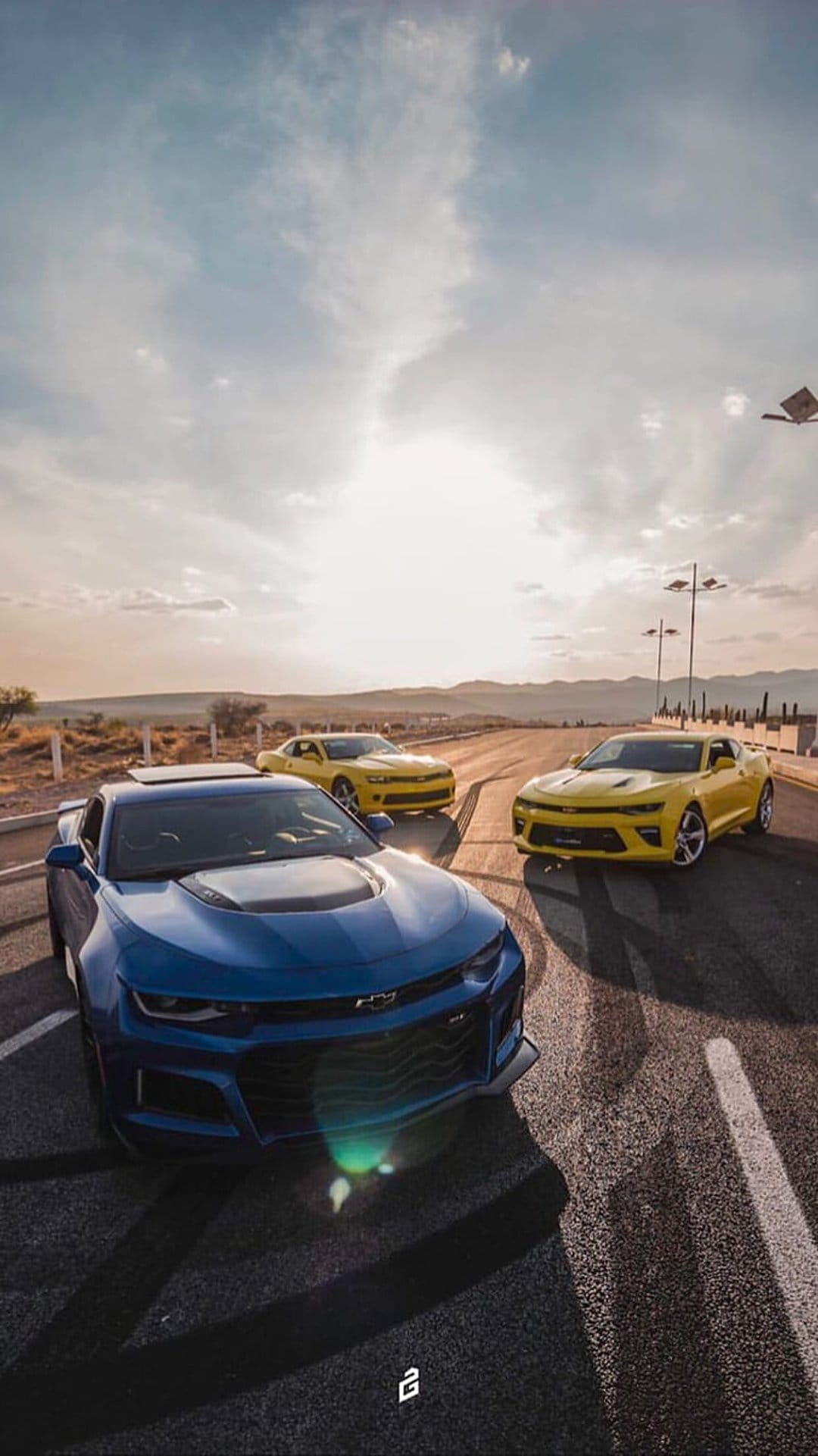 Three Chevrolet Camaros, one blue and two yellow, on a desert road with skid marks at sunset under a warm sky.