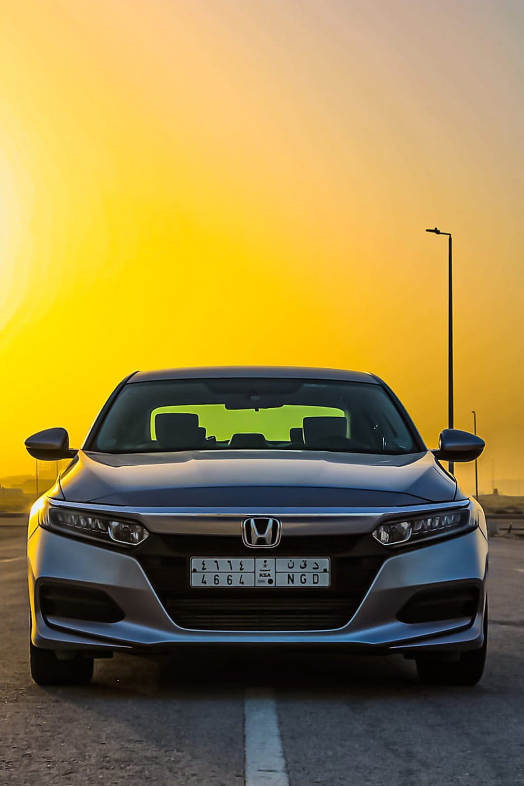 Silver Honda Accord sedan with glowing headlights faces forward against a golden orange sunset sky and street lamp.