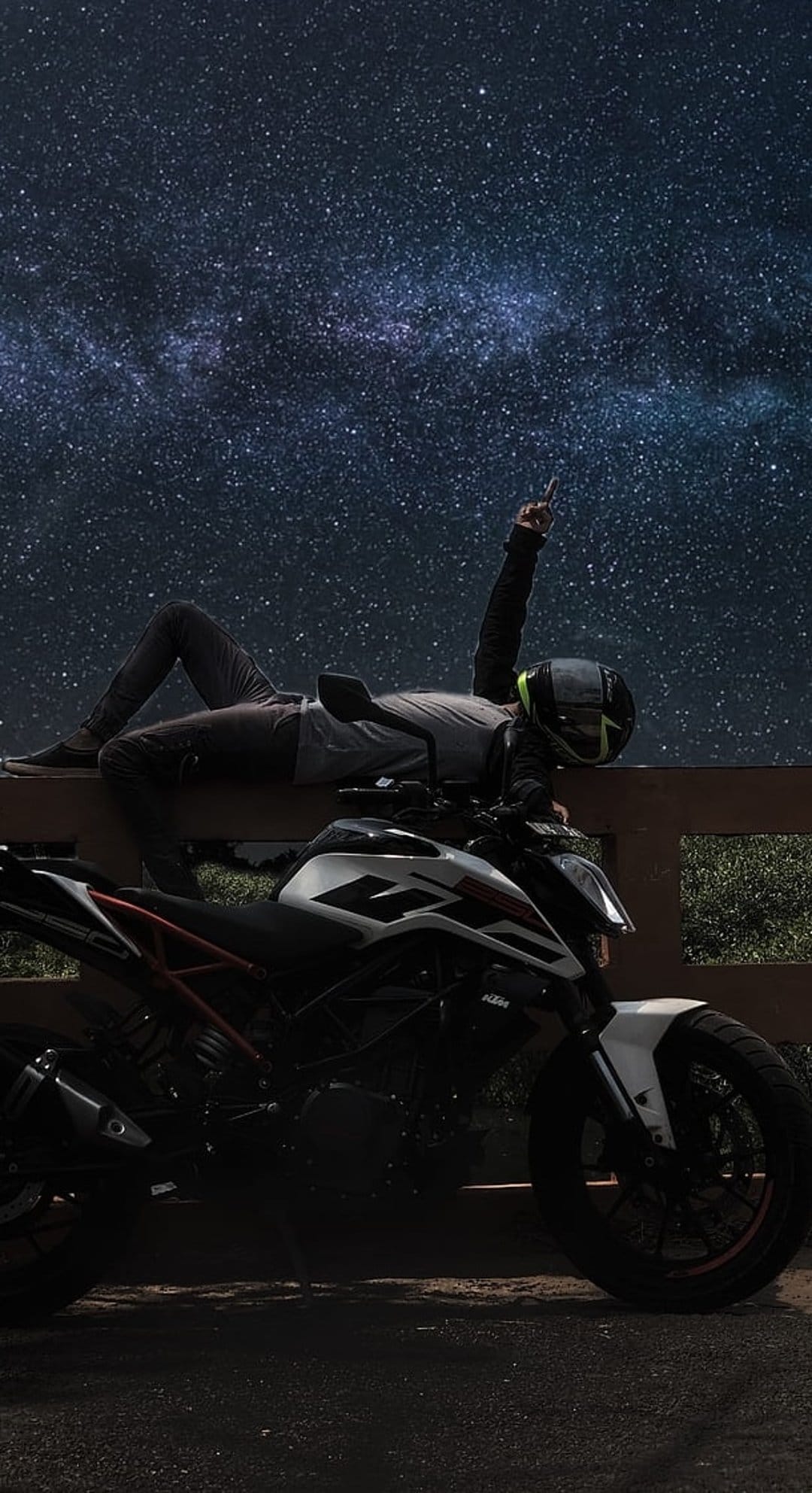 Motorcyclist in gear reclining on a wooden rail beneath a vibrant Milky Way galaxy and a parked bike at night.