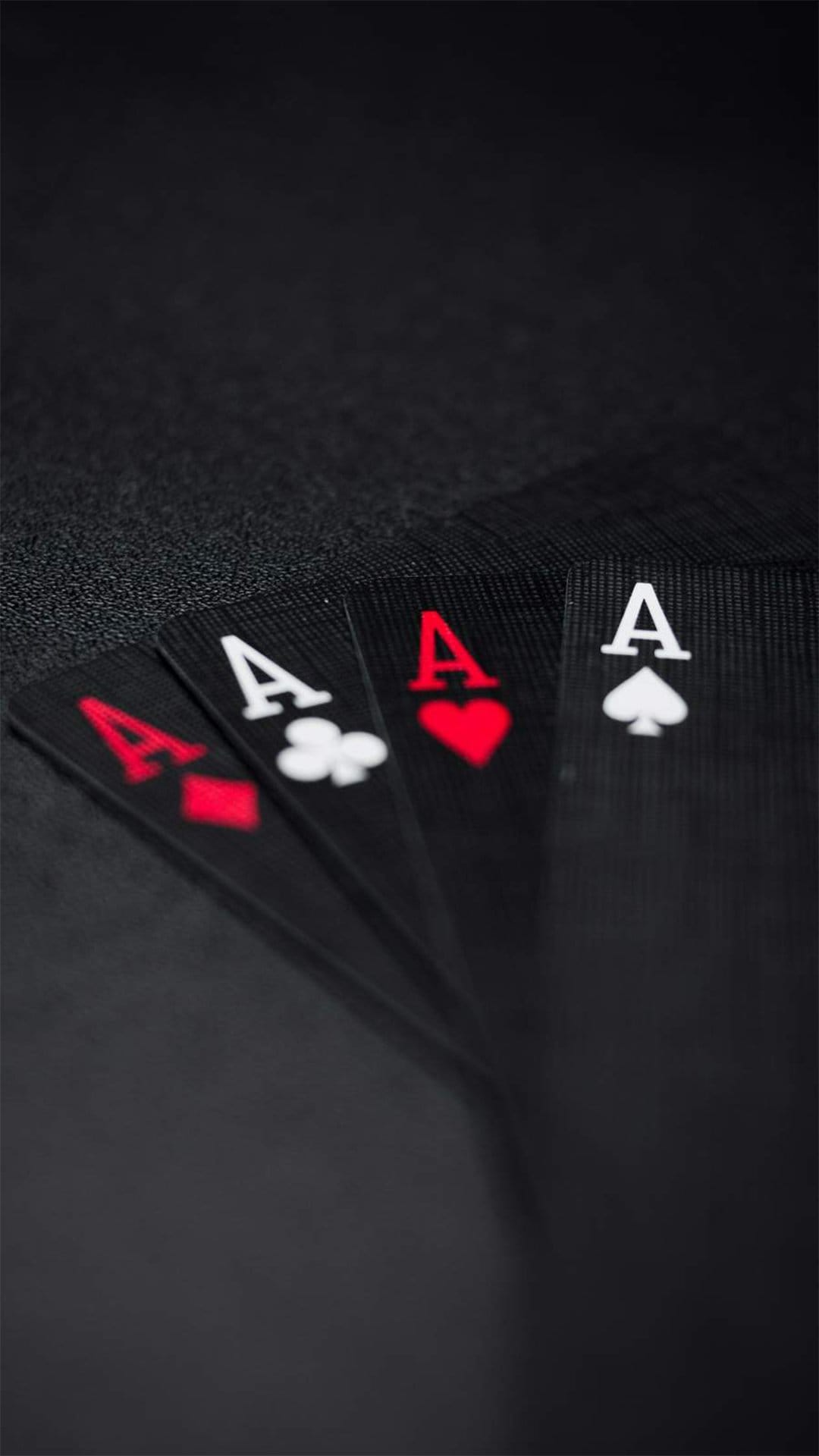 Four fanned ace playing cards rest against a dark textured surface with subtle red and black suit icons visible.