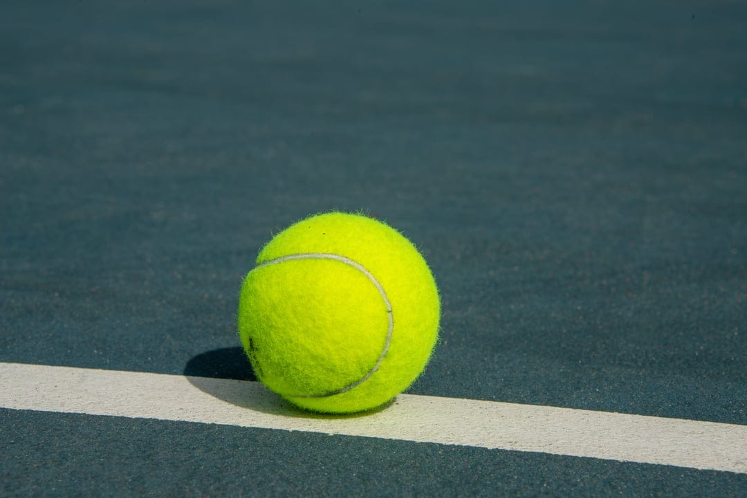Yellow tennis ball resting on a textured blue hard court surface next to a solid white boundary line.