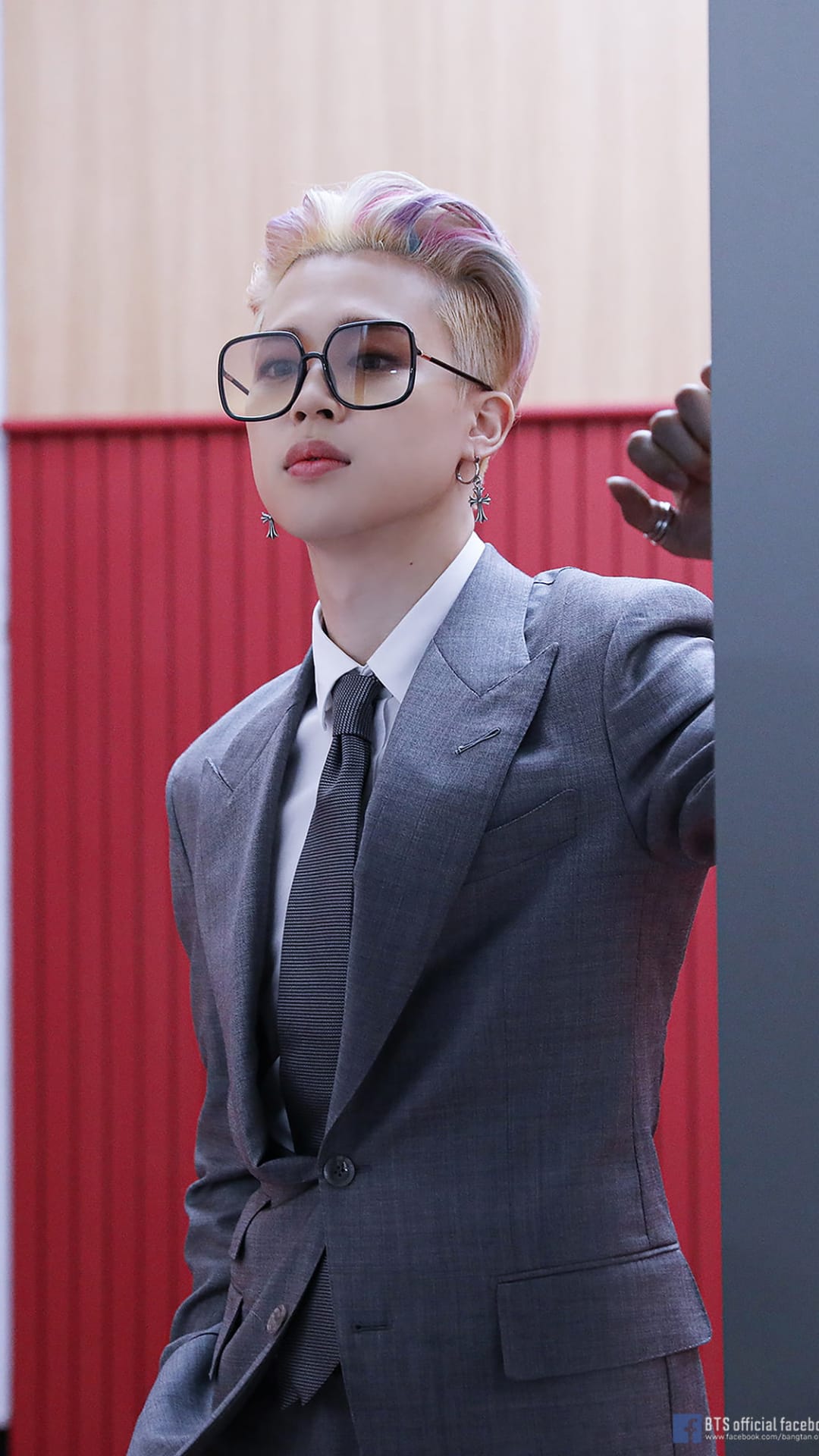 Jimin of BTS wearing a pinstripe suit and tinted square glasses against a red and grey textured background wall.