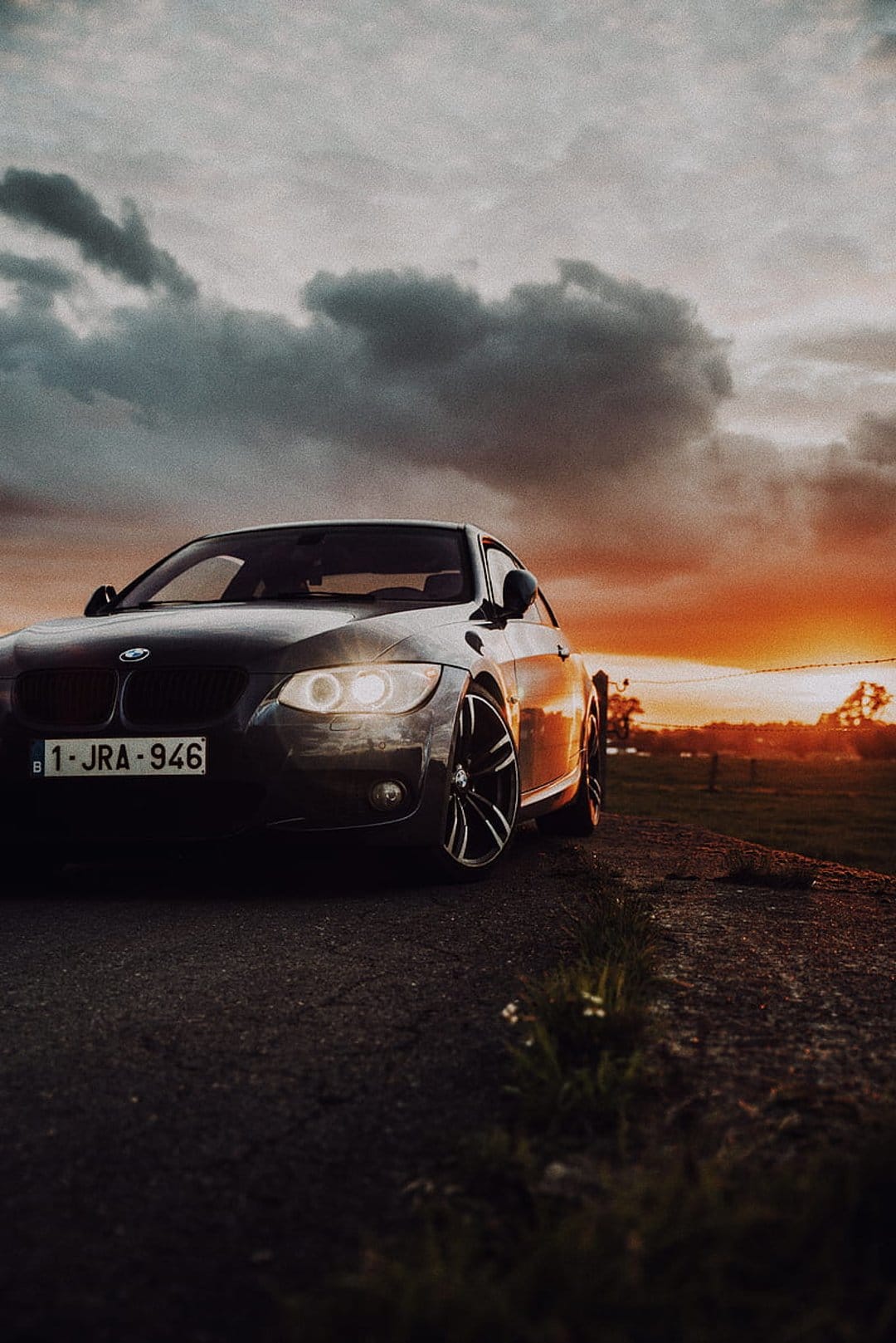 Dark BMW coupe with lit headlights on a gravel road under a dramatic orange and purple sunset sky with clouds.