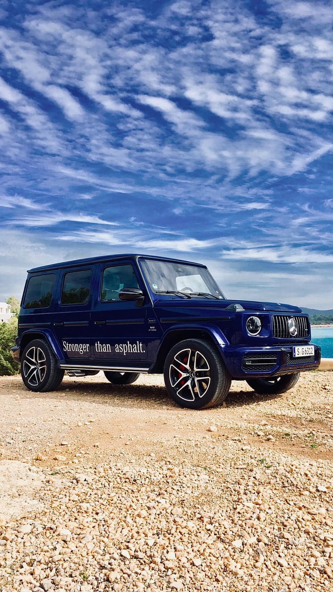 Navy blue Mercedes G-Wagon SUV parked on coastal sands under a clear sky with 'Stronger than asphalt' text.