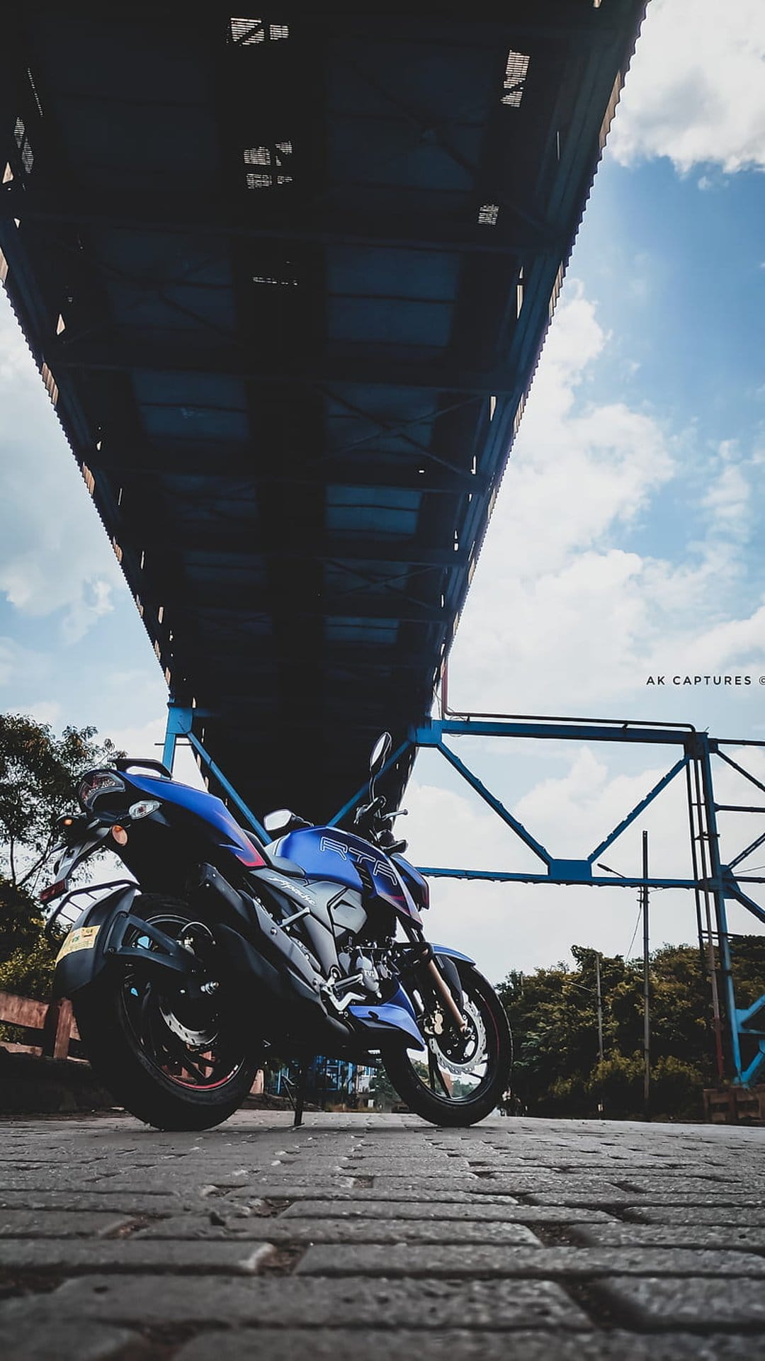 Blue sportbike on cobblestone road beneath a massive bridge structure with a bright cloudy sky background.