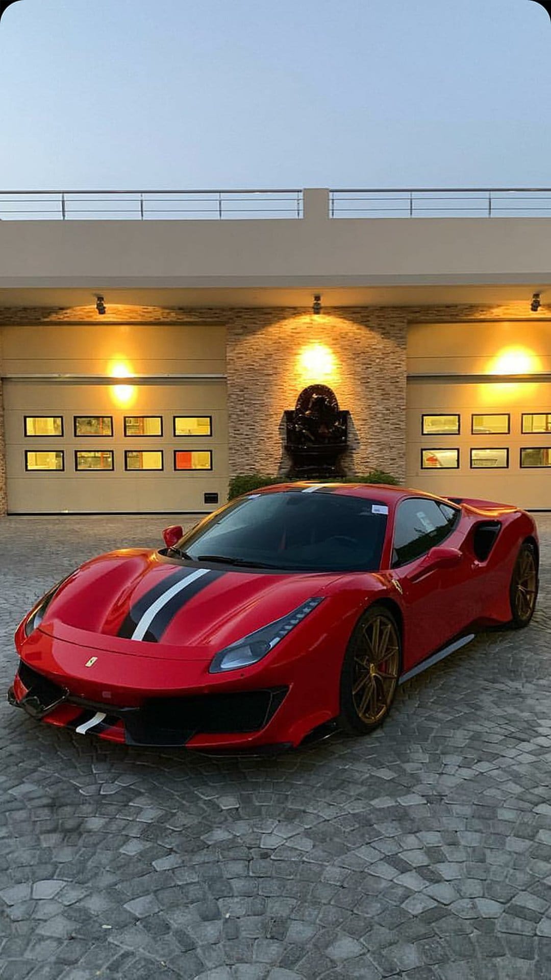 Red Ferrari 488 GTB with racing stripes parked on a cobblestone driveway in front of a modern lighted garage.