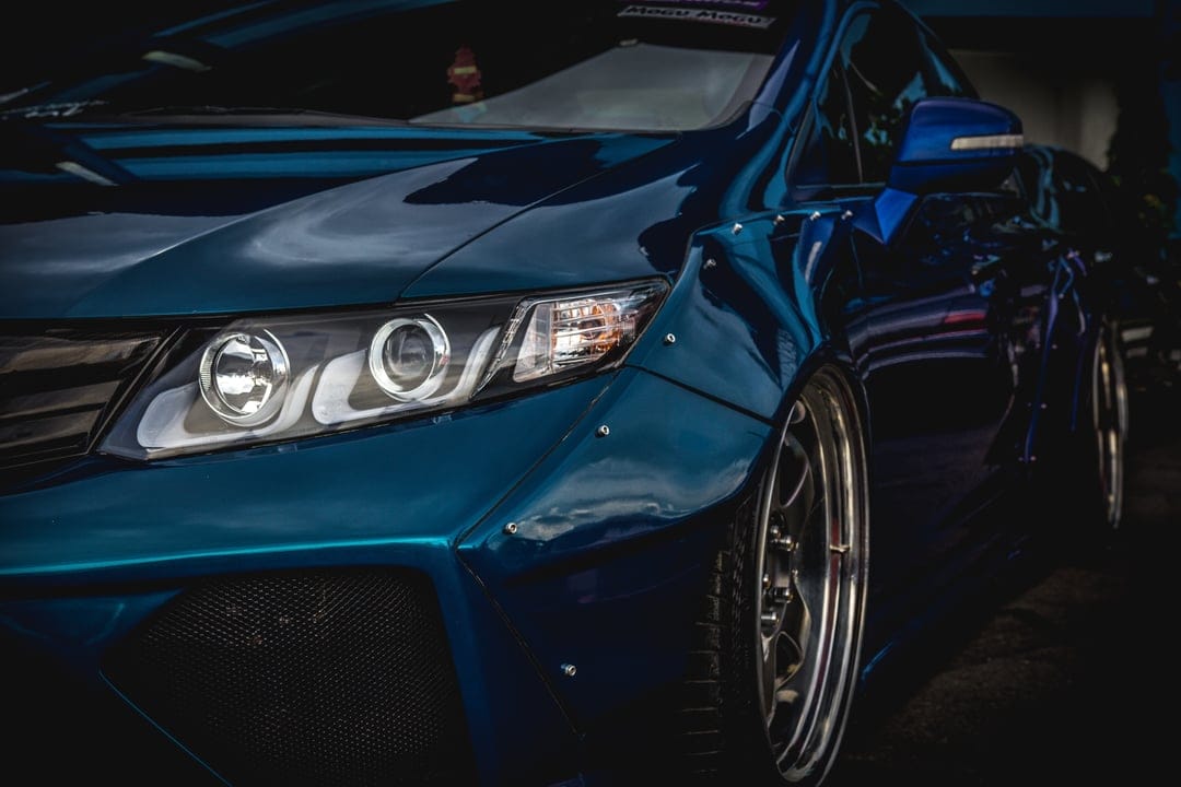 Metallic blue sports car with flared widebody fenders and glowing circular LED headlights in a low-angle shot.