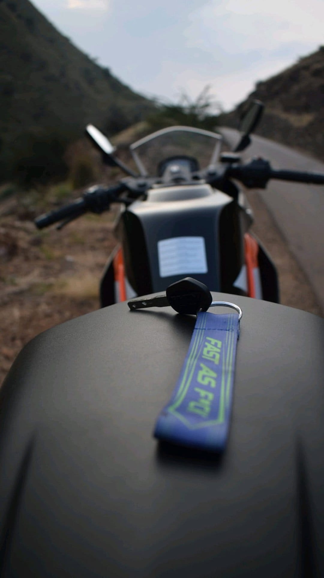 KTM motorcycle keys with a blue keychain on a black seat with misty mountains and a cloudy sky behind.