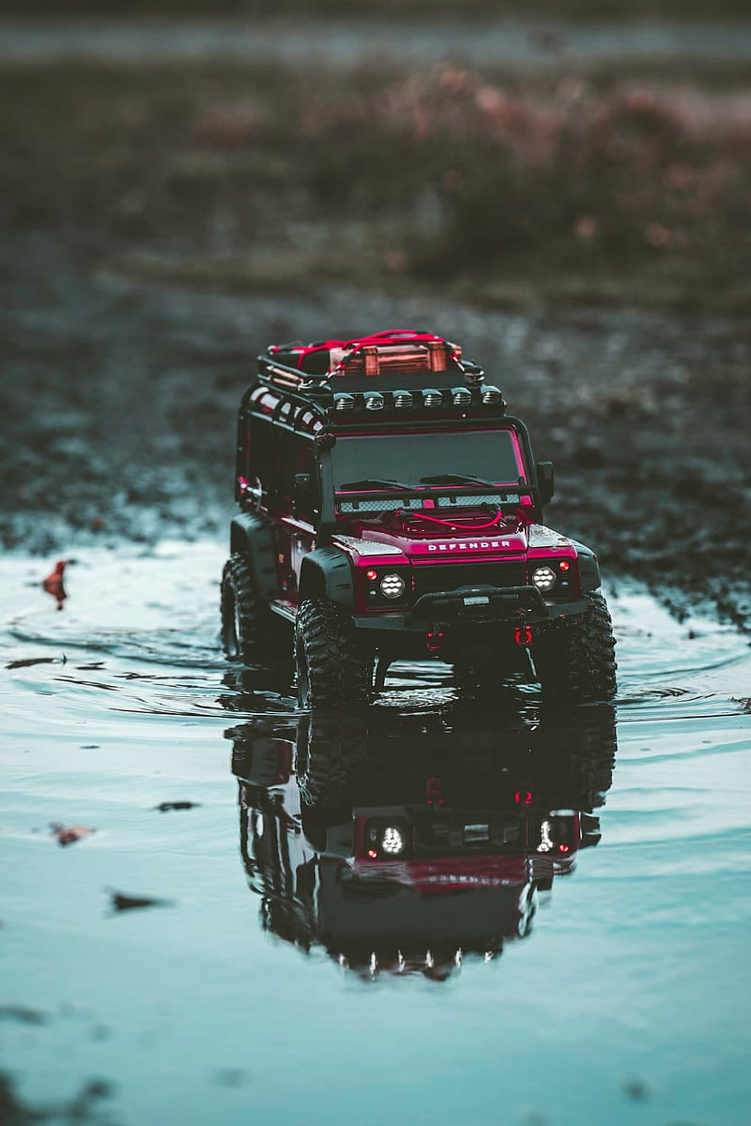 Red scale model SUV with roof rack and off-road tires reflects in a shallow water puddle under moody lighting.