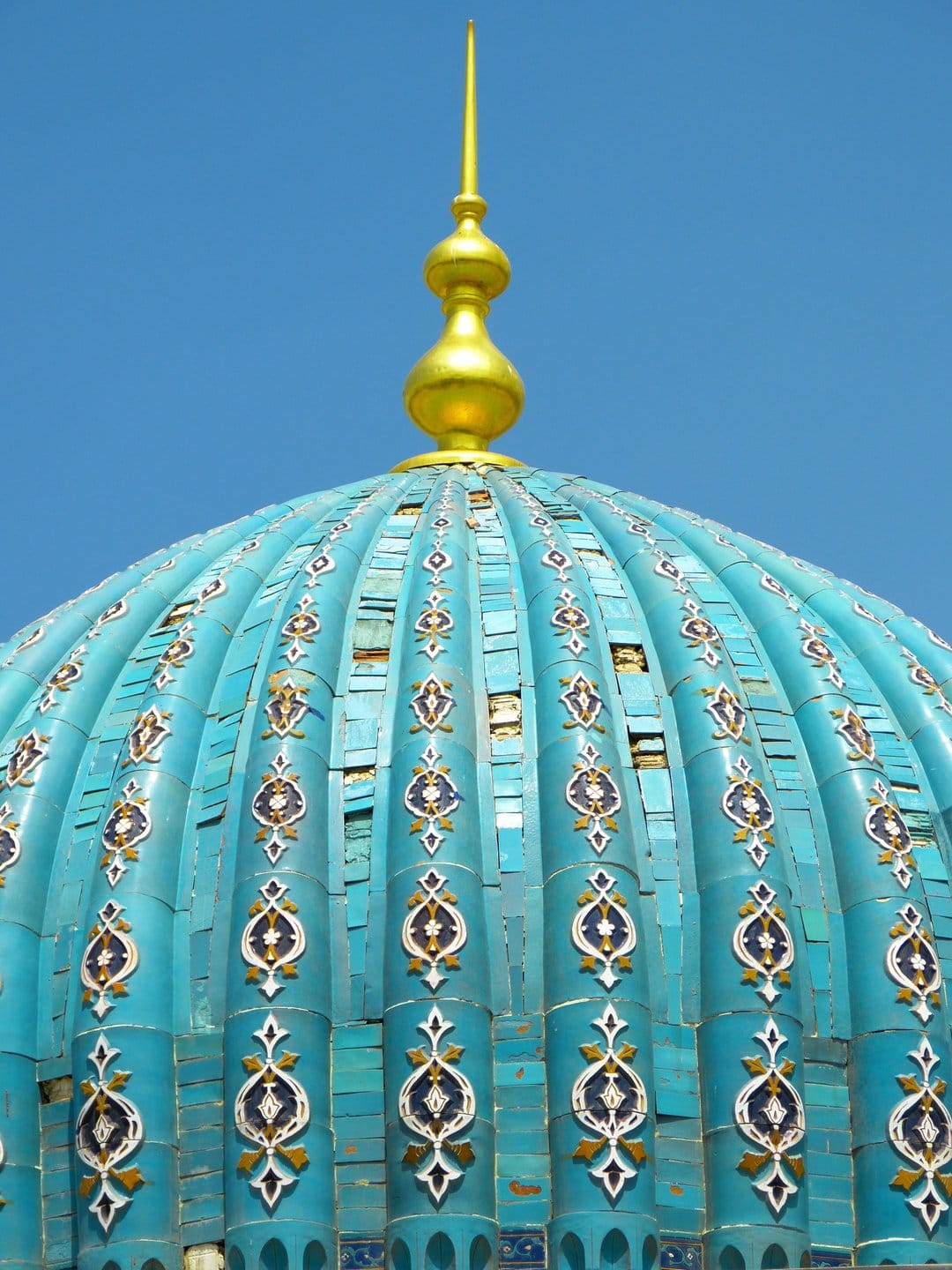 Turquoise mosaic dome with a golden spire and floral patterns against a clear blue sky in vertical orientation.