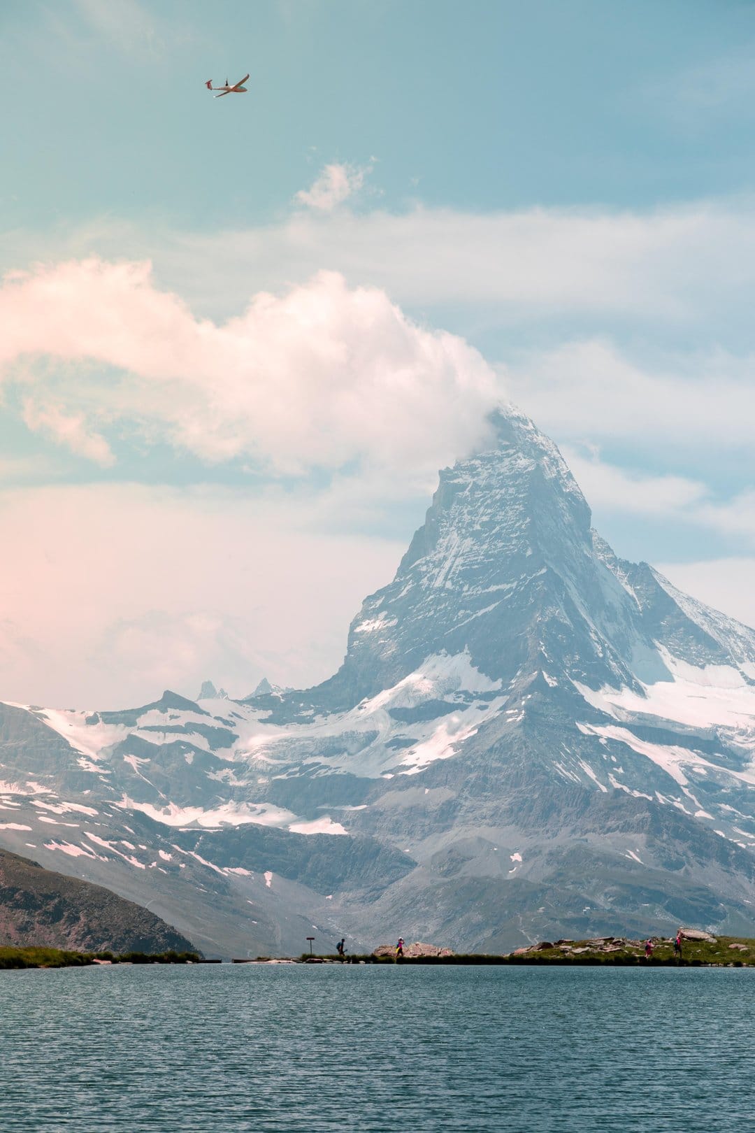 Snow-capped Matterhorn mountain reflects in a still lake with a small airplane flying through a pale blue sky.