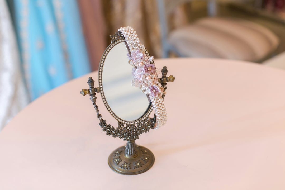 Antique oval vanity mirror draped with a pearl and pastel floral headband resting on a soft pink tabletop surface.
