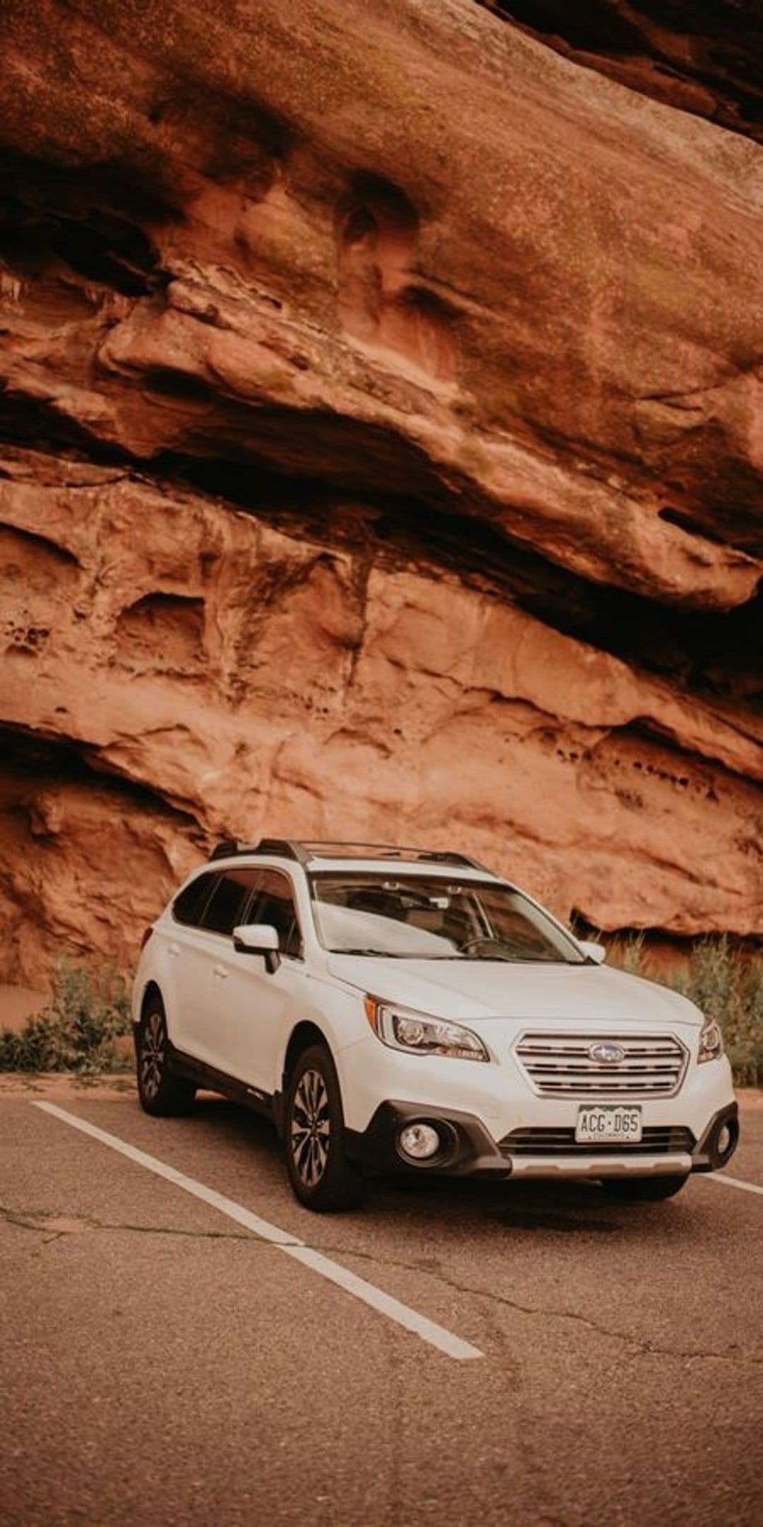 White Subaru Outback parked at the base of a massive, textured red sandstone cliff in a desert landscape.